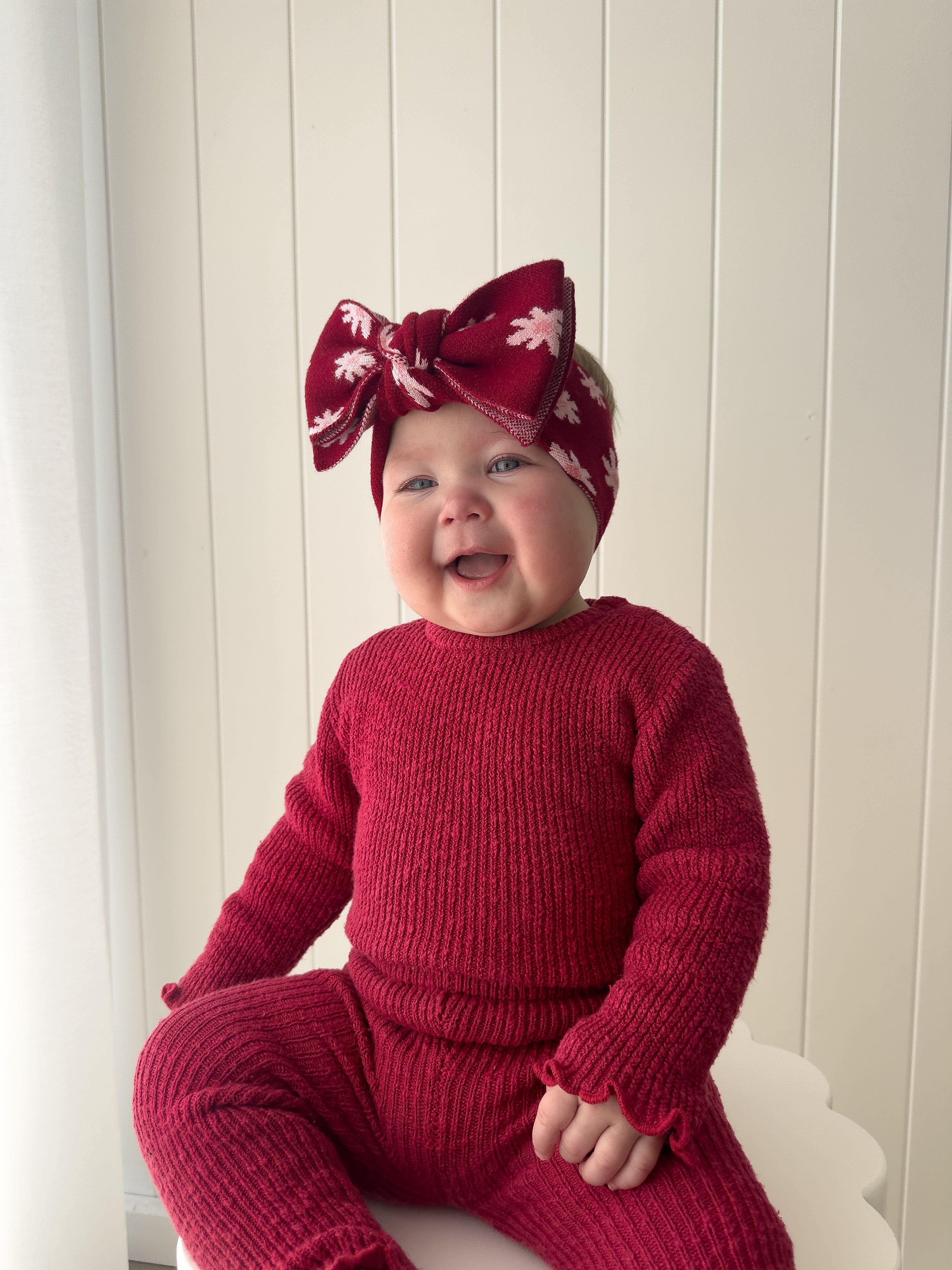 A smiling baby in a matching red knit outfit and a Ziggy Lou - Headband Lola (Plum) by ZIGGY LOU sits against a white paneled wall.