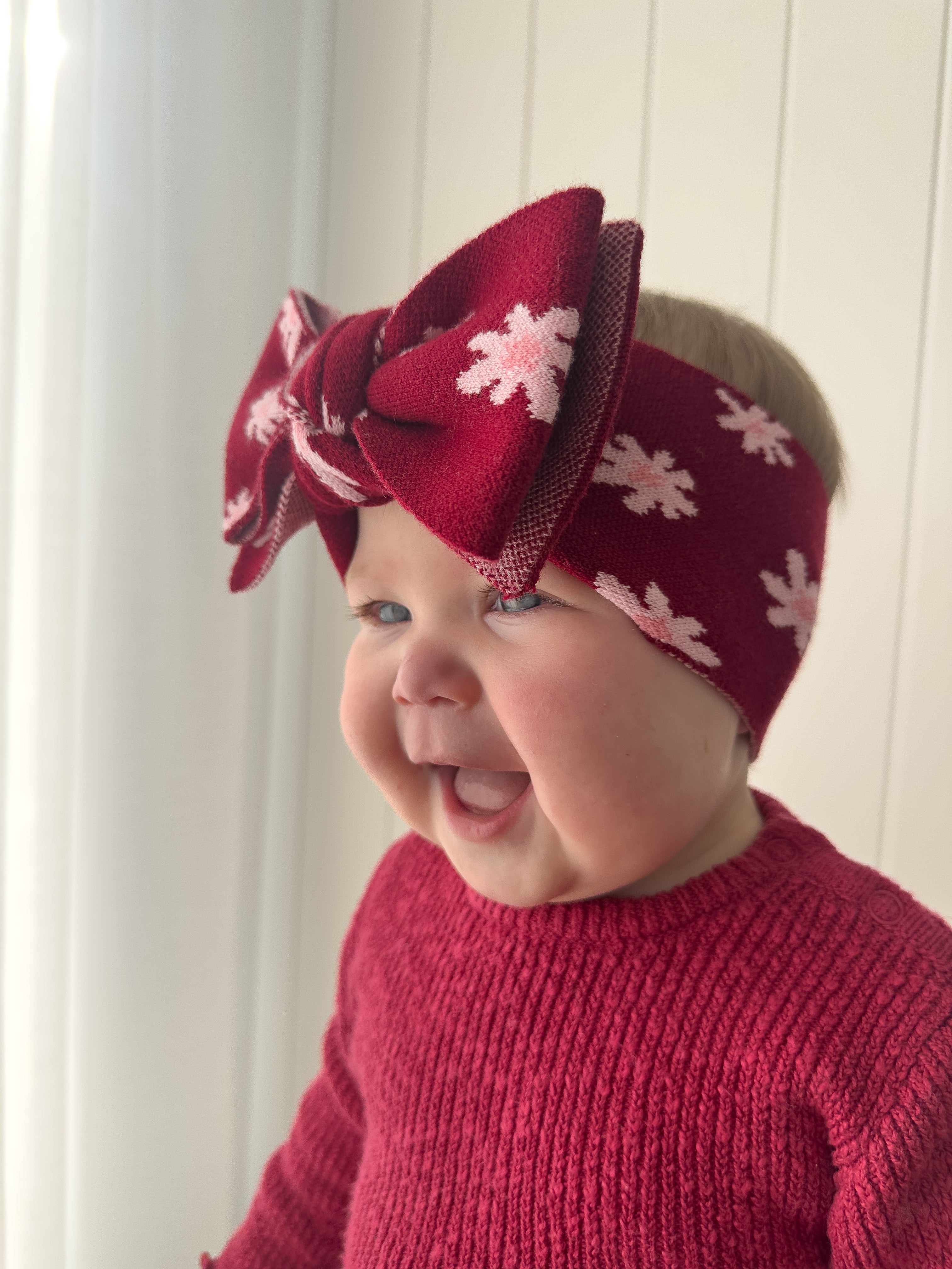 A smiling baby wears a red knit sweater and a ZIGGY LOU Headband Lola (Plum) with a large bow, standing in front of a white wall and curtain.