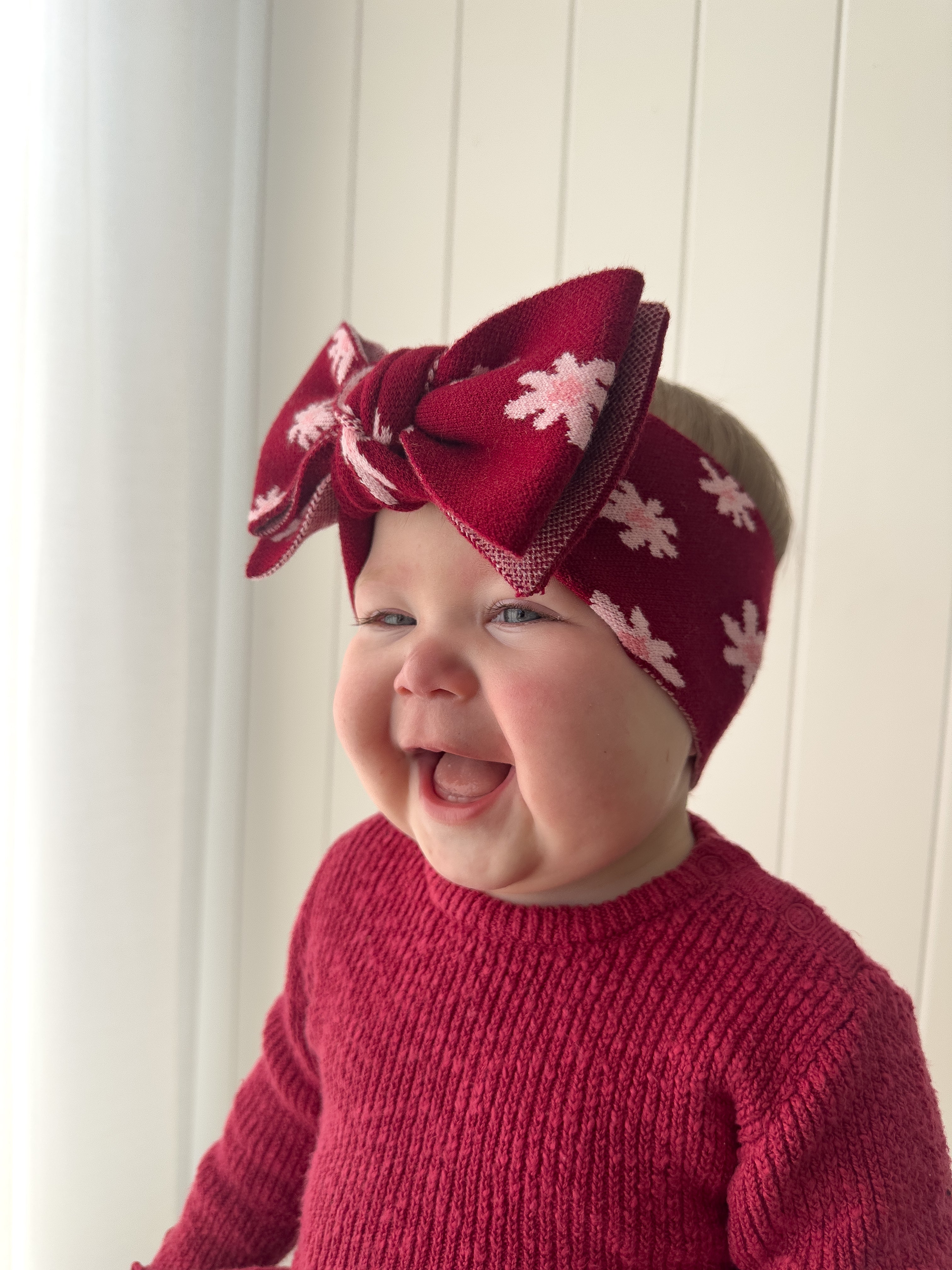 A smiling baby in a red knit sweater wears the ZIGGY LOU Ziggy Lou - Headband Lola (Plum), featuring a large bow, against a white paneled background.