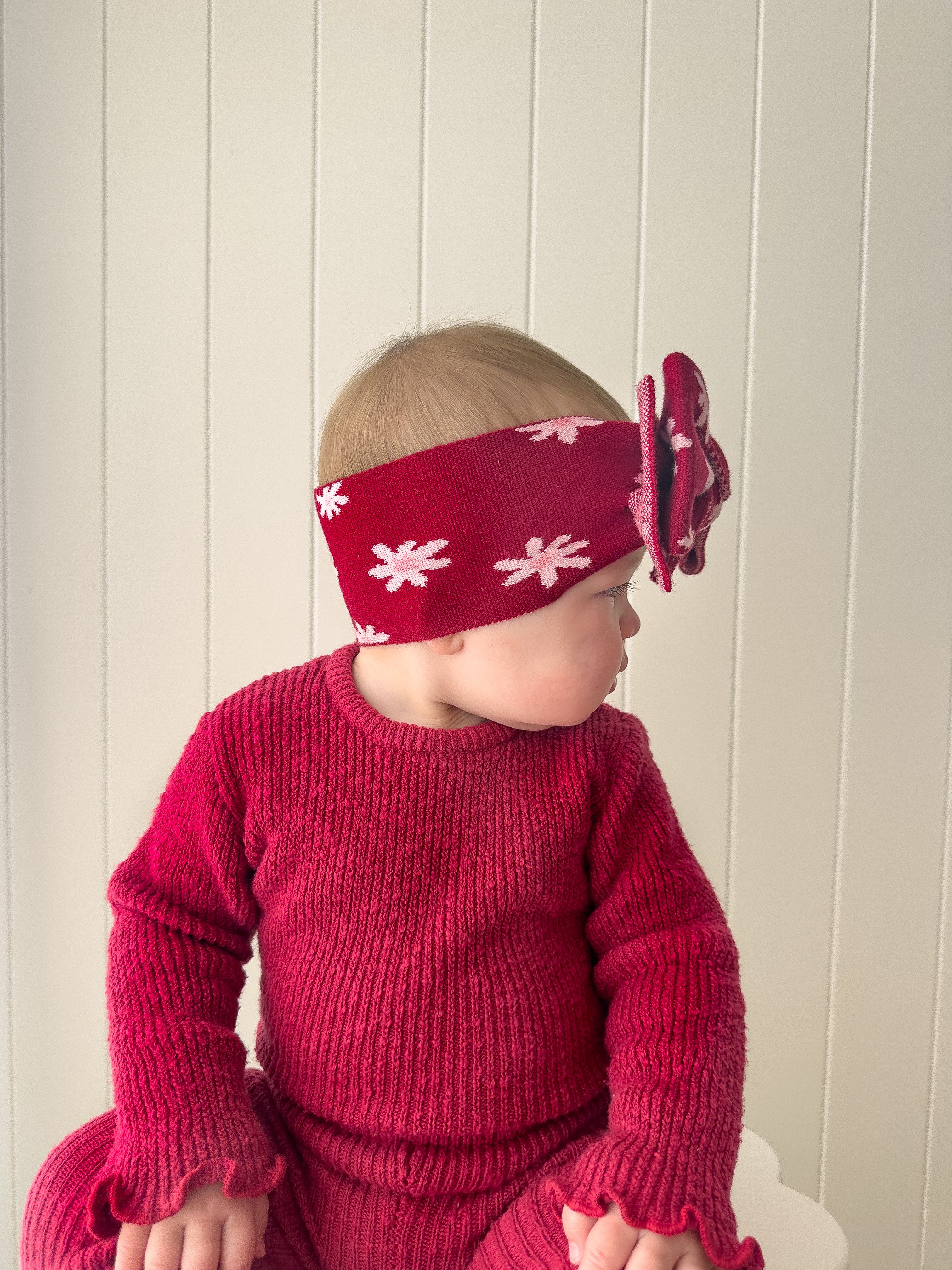 A baby in a red knit outfit wears the ZIGGY LOU Ziggy Lou - Headband Lola (Plum), featuring a large bow, while looking to the side against a light-paneled background.