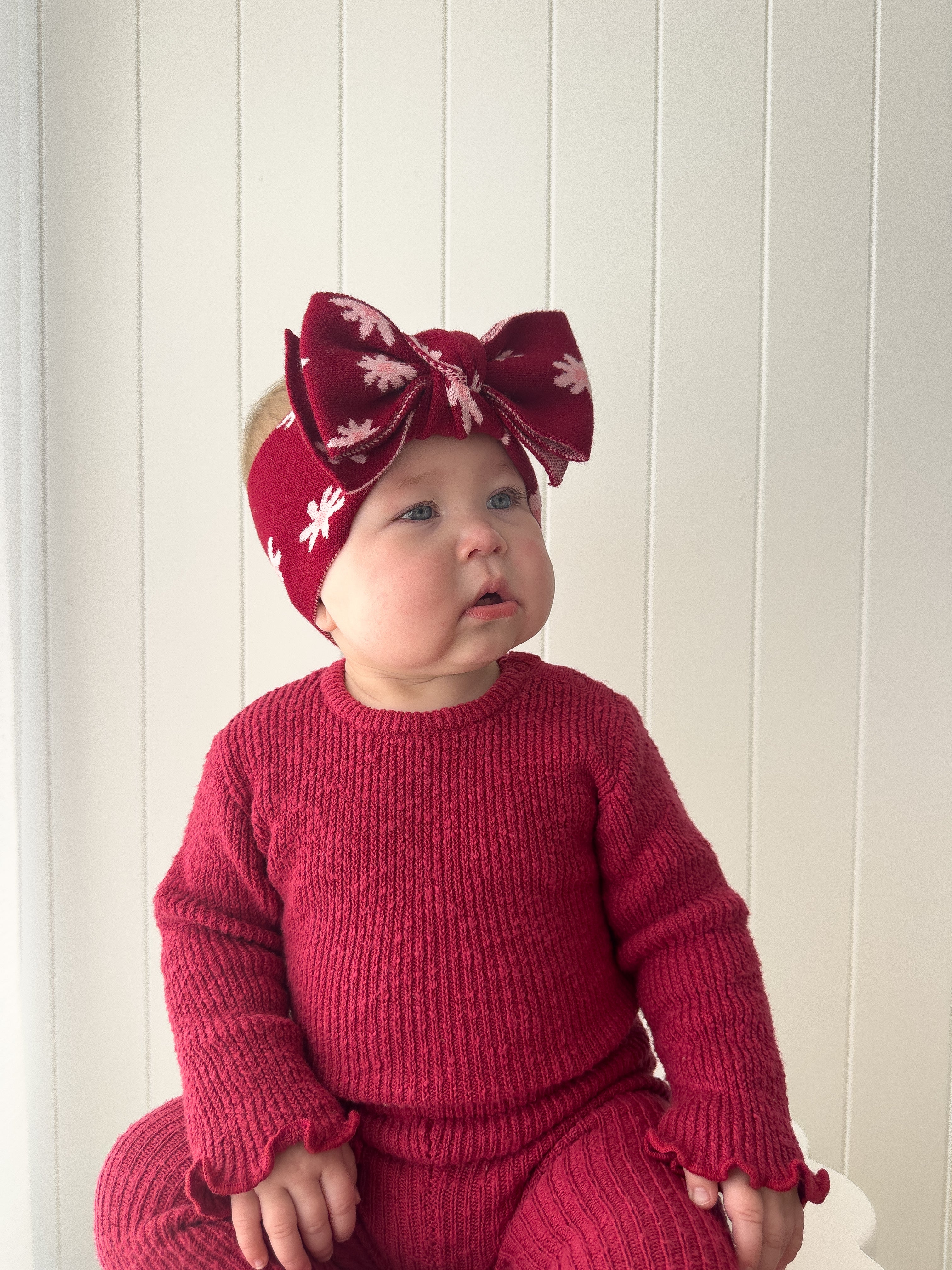 A baby wears the ZIGGY LOU - Headband Lola (Plum) and a red knit outfit, sitting against a white paneled wall and looking slightly to the side.
