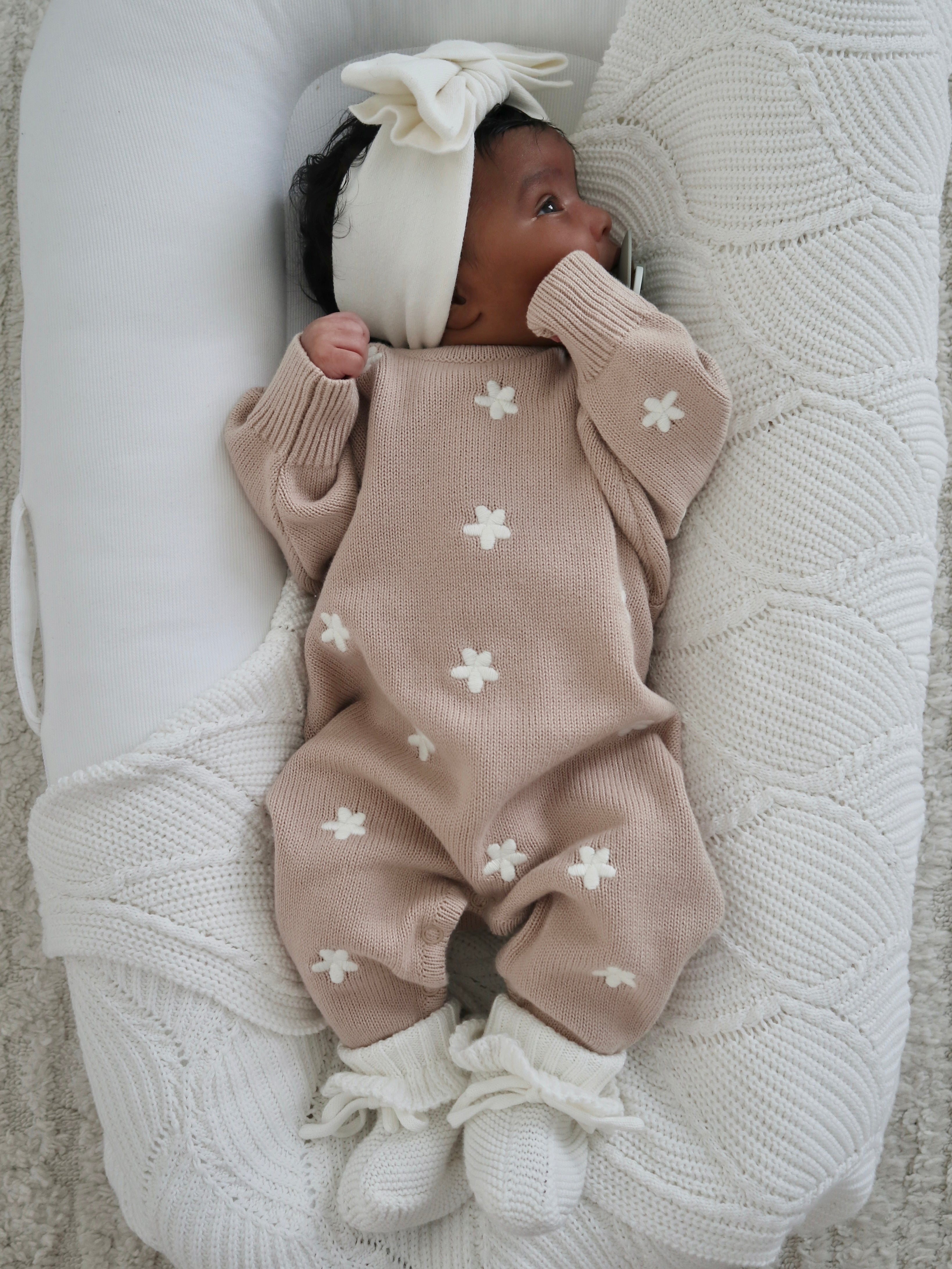 A baby in a ZIGGY LOU Winter Playsuit Aurora (Size NB)—a beige knit with white flowers—and a white headband rests in a cream-colored, textured baby nest on a soft, light backdrop.