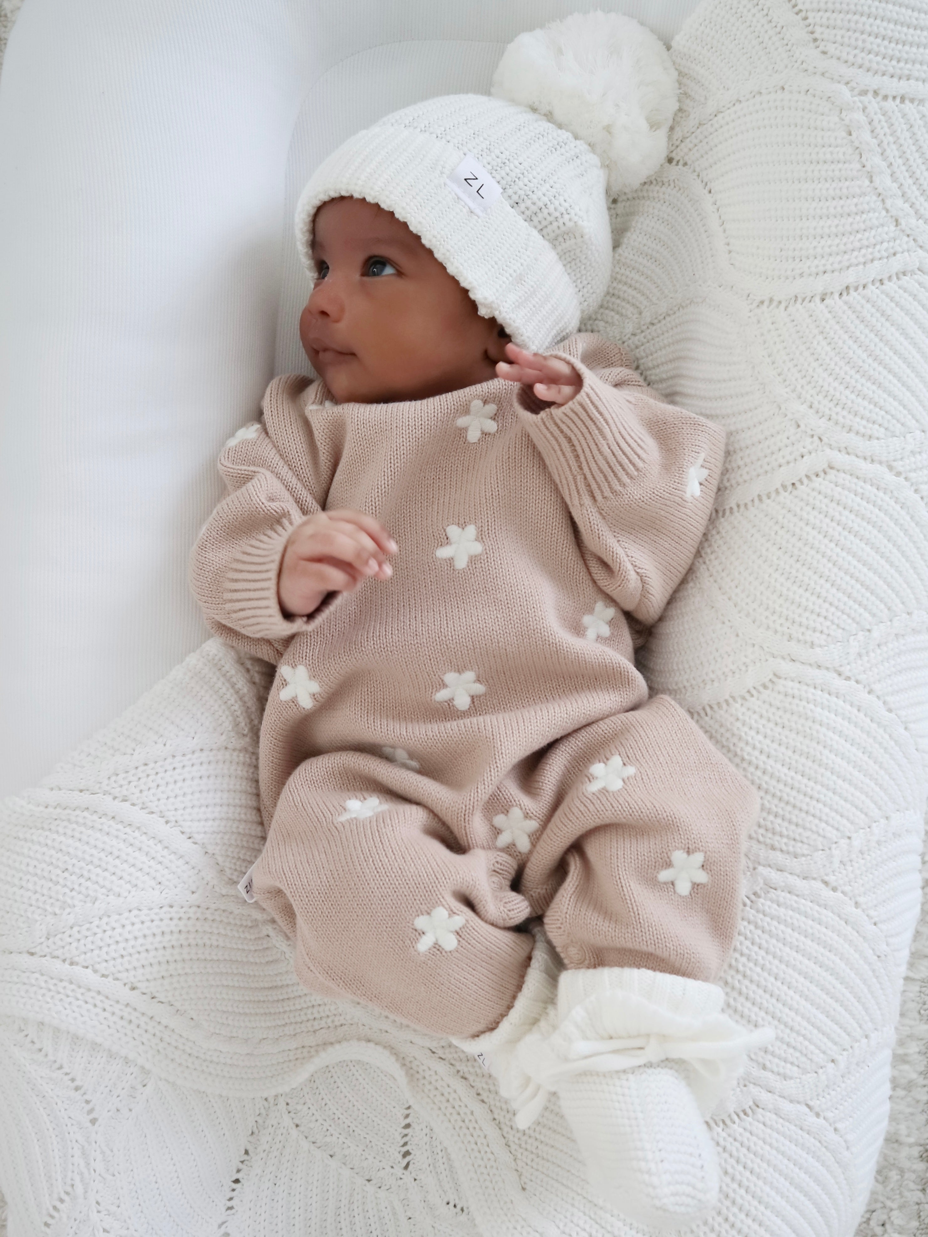 A baby lies on a white knitted blanket, wearing the ZIGGY LOU Winter Playsuit Aurora in size NB with white flower patterns and a matching knit hat with pom-pom, looking up with hands slightly raised.