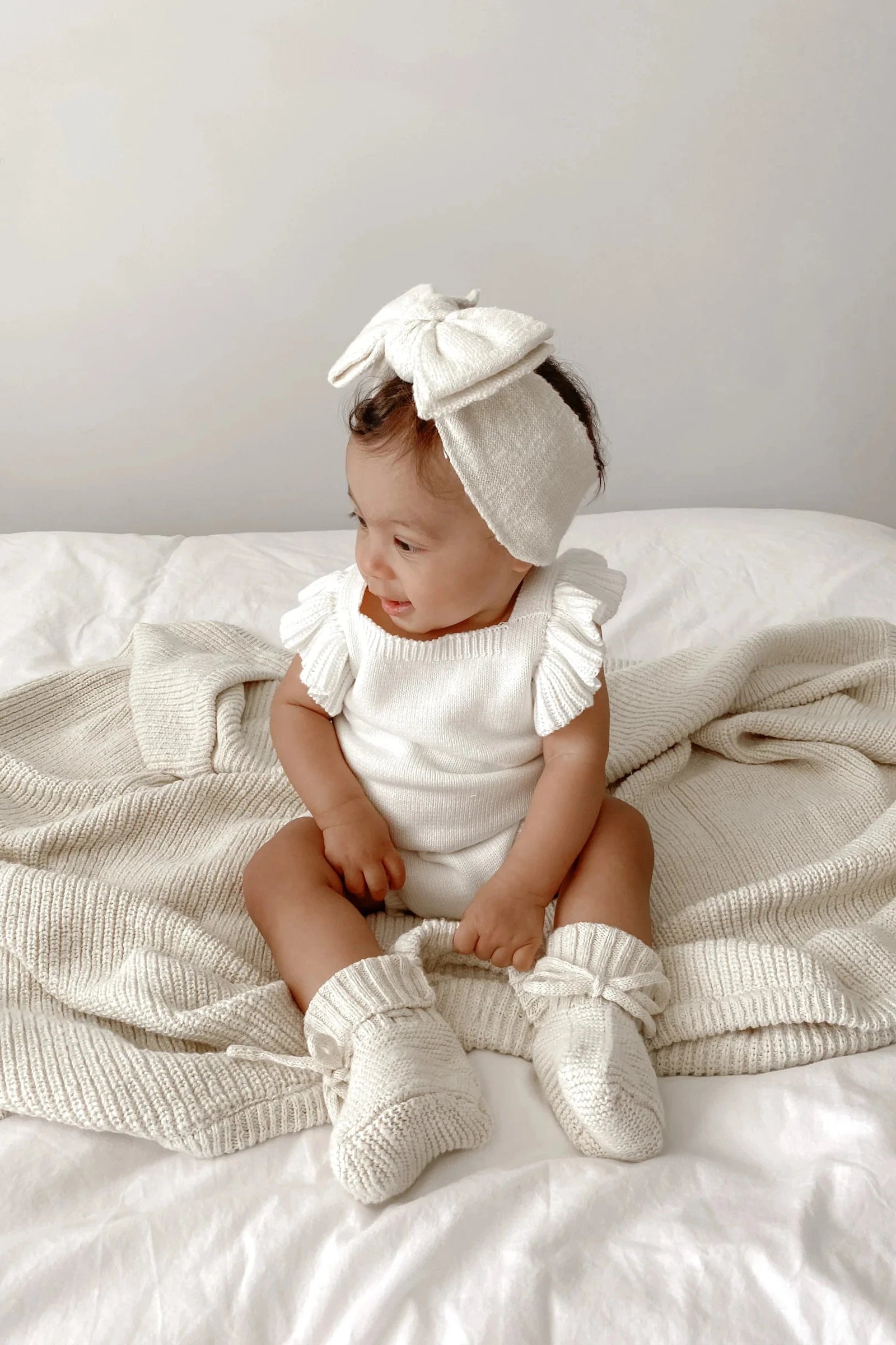A baby sits on a bed with a cream knit blanket, wearing a white outfit with ruffled sleeves, a large bow headband, and ZIGGY LOU - Booties Honey. The baby smiles while looking to the side.