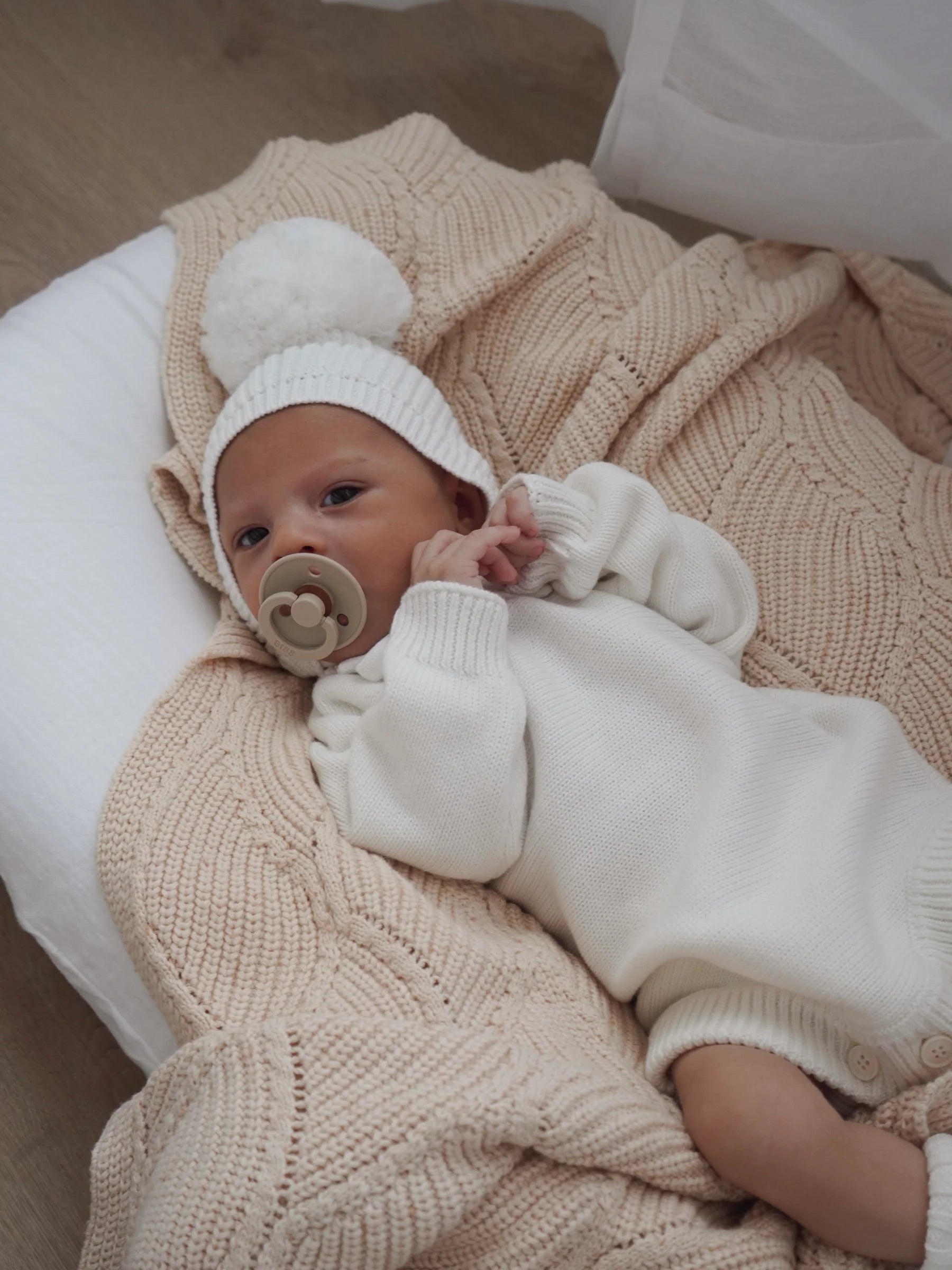 A baby in a white cotton knit outfit and pom-pom hat lies calmly on the ZIGGY LOU Ziggy Lou - Blanket Biscotti Fleck, holding a pacifier and gazing upward.