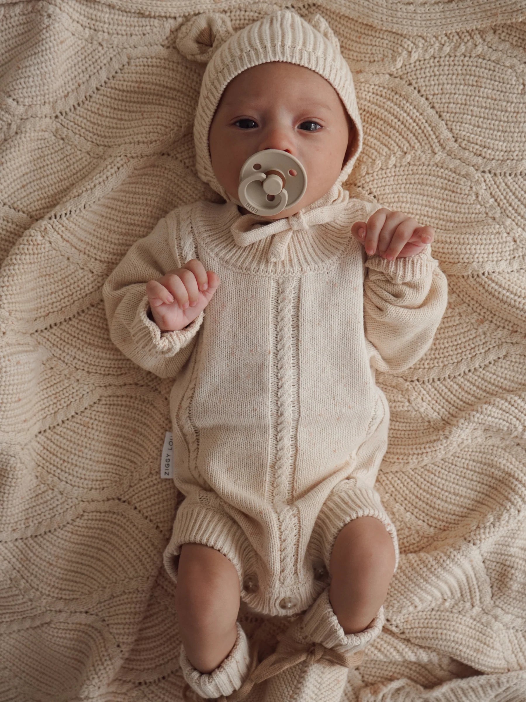 A baby with a pacifier, dressed in a beige knit outfit with hood and booties, lies on the ZIGGY LOU Ziggy Lou - Blanket Biscotti Fleck, looking up at the camera.
