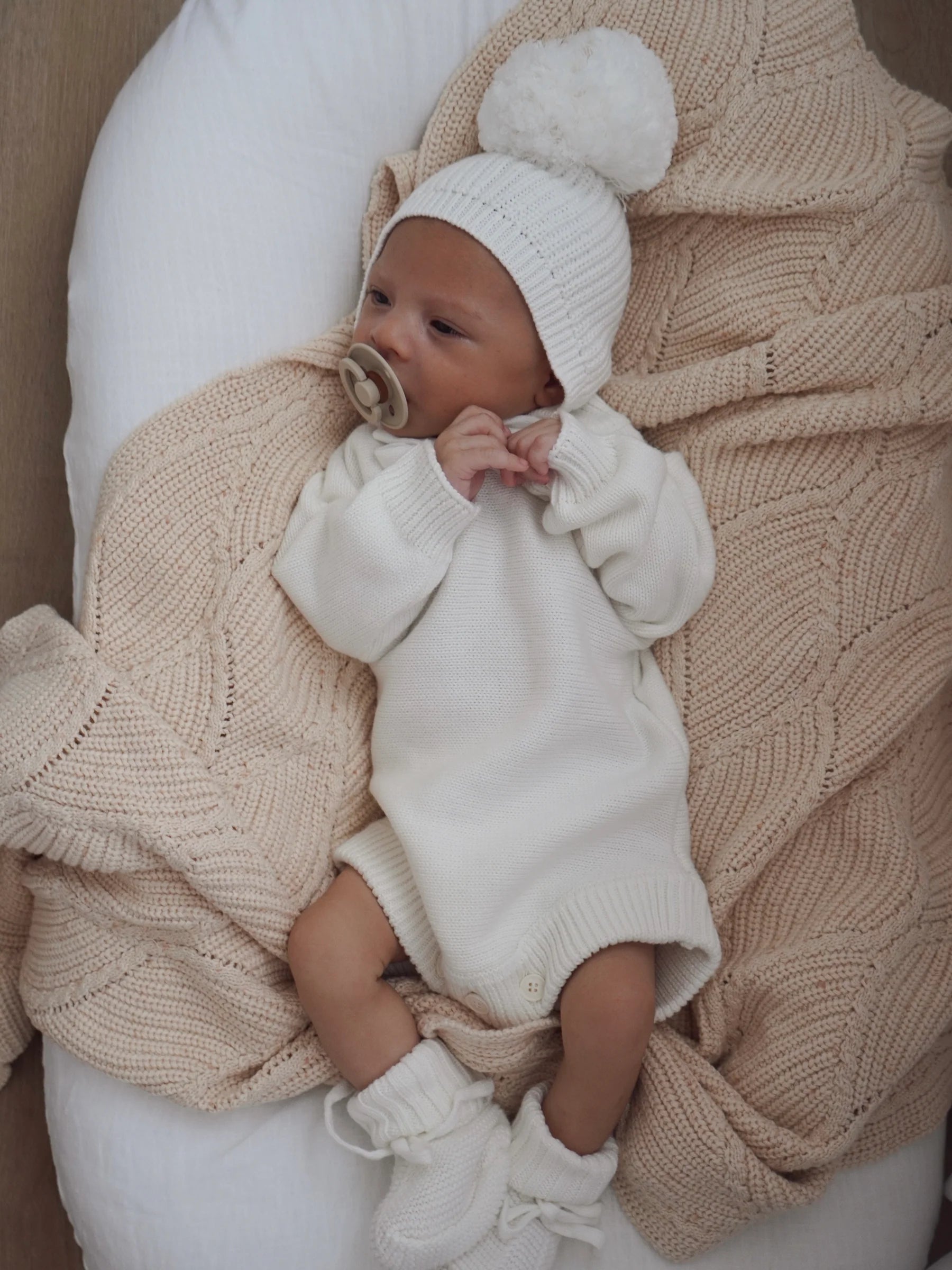 A baby lies on a cream knit blanket, wearing the ZIGGY LOU Ziggy Lou - Beanie Milk from the Milk Signatures Collection, a white onesie, matching 100% cotton booties, and a pacifier. The baby looks to the side with hands near their face.