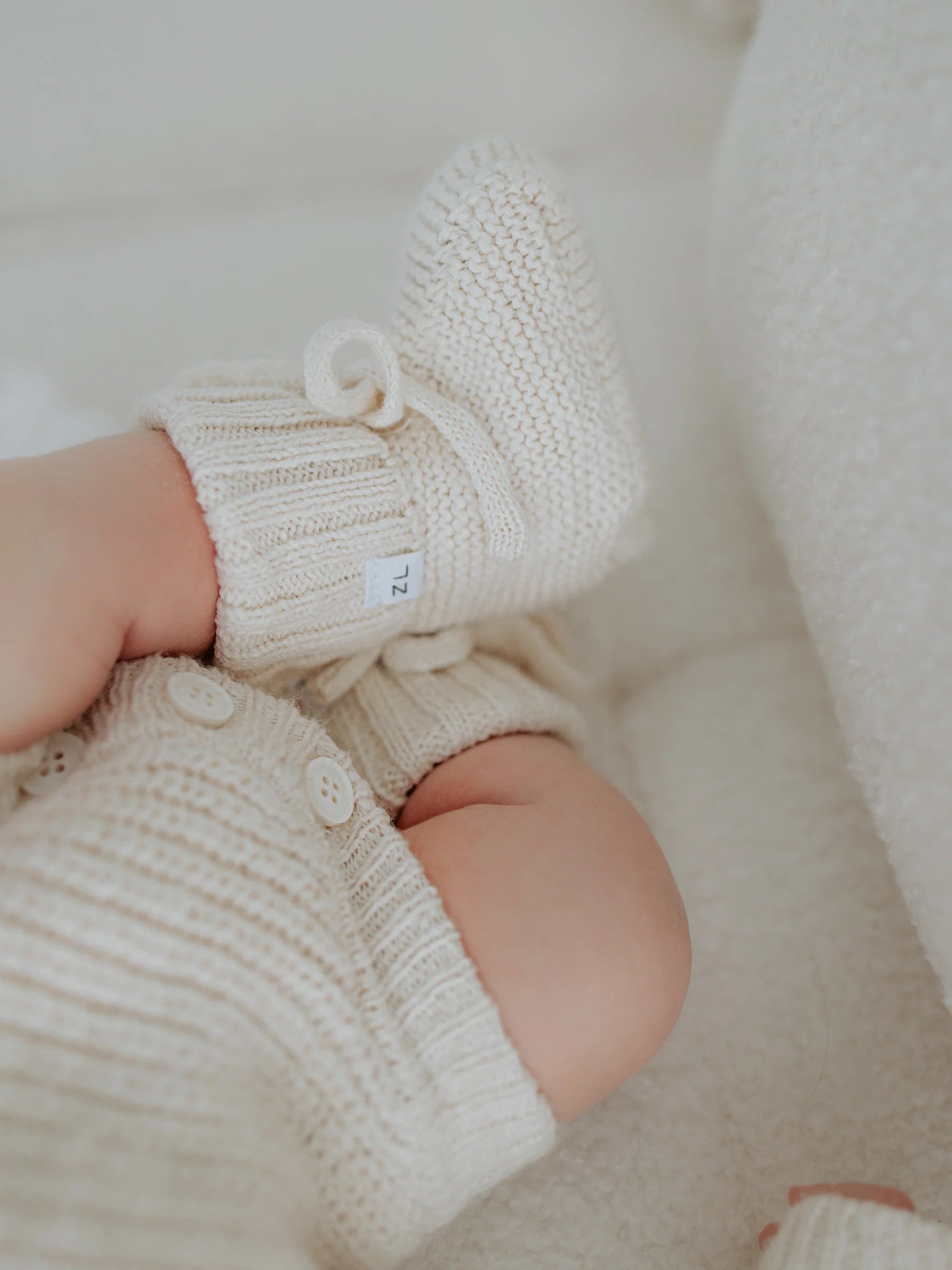 A close-up of a baby's legs wearing ZIGGY LOU’s Ziggy Lou - Booties Honey and matching knit shorts with buttons, lying on a soft textured surface—the perfect heirloom keepsake.