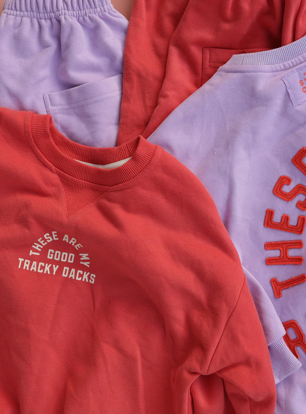 Red and lavender sweatshirts and shorts are displayed. The SMALL SWIM CLUB Good Tracky Pullover in red/cream reads "THESE ARE MY GOOD TRACKY DACKS" in white, while the vintage fleece lavender set features partially visible red text.