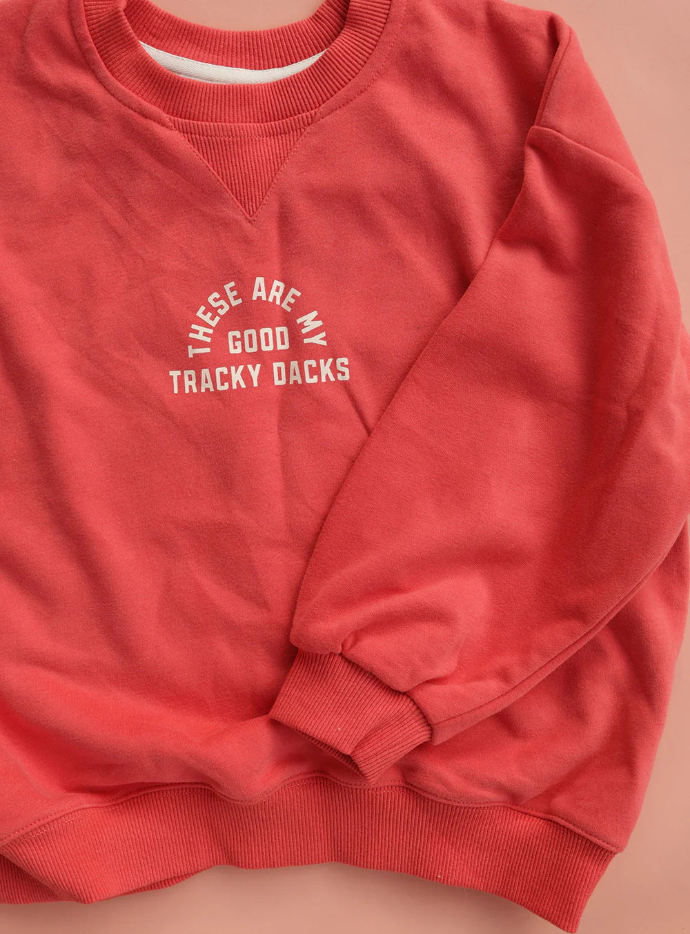 The SMALL SWIM CLUB Small Swim Club - Good Tracky Pullover Red/Cream features a slouchy fit and “THESE ARE MY GOOD TRACKY DACKS” in white arching across the chest, shown flat on a peach background.