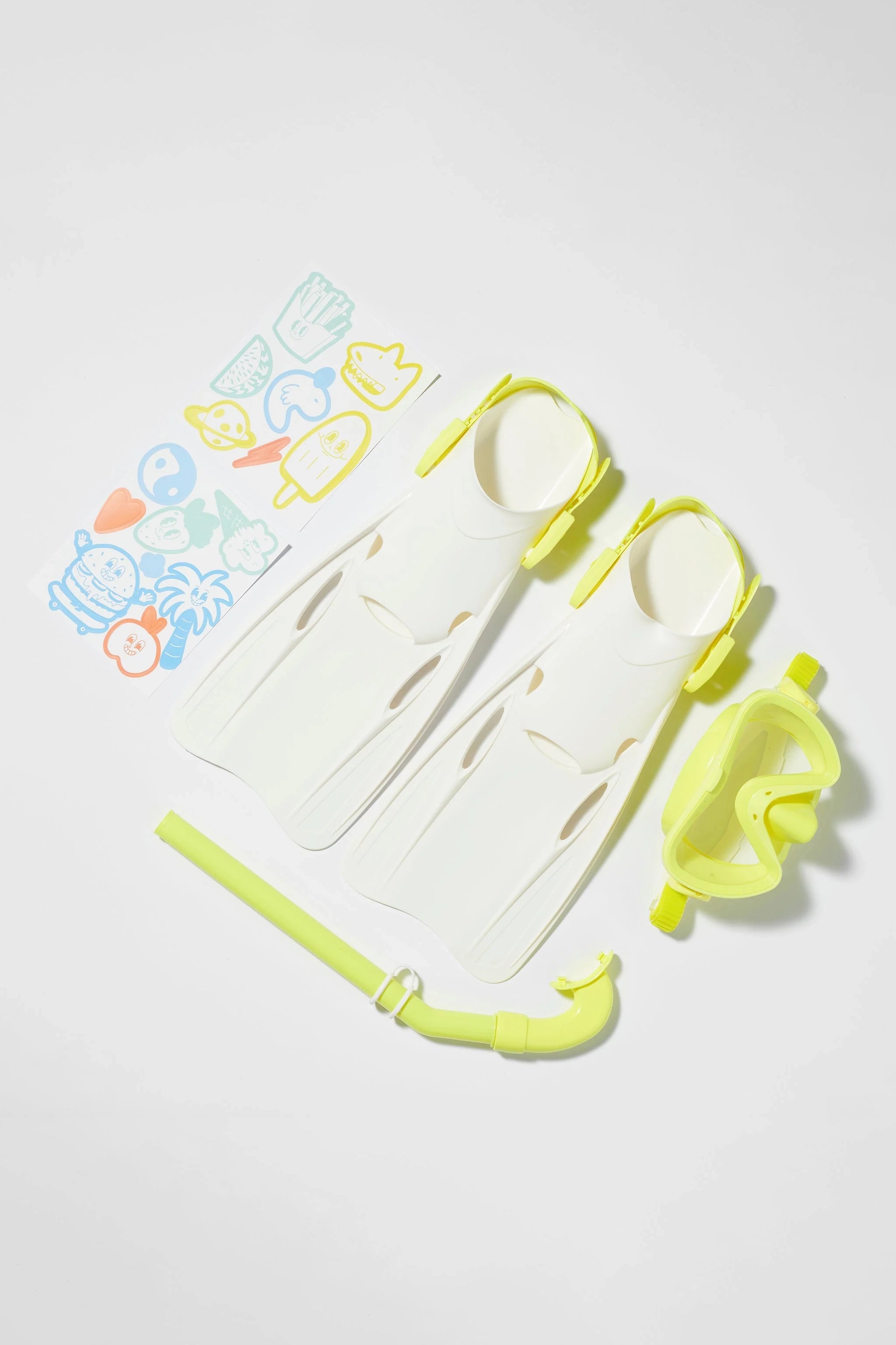 A set of the SUNNYLIFE Kids Snorkel Set Yellow, featuring flippers, a snorkel, and a mask, is laid out on a white background. Colorful tropical-themed stickers are arranged to the left of the gear.