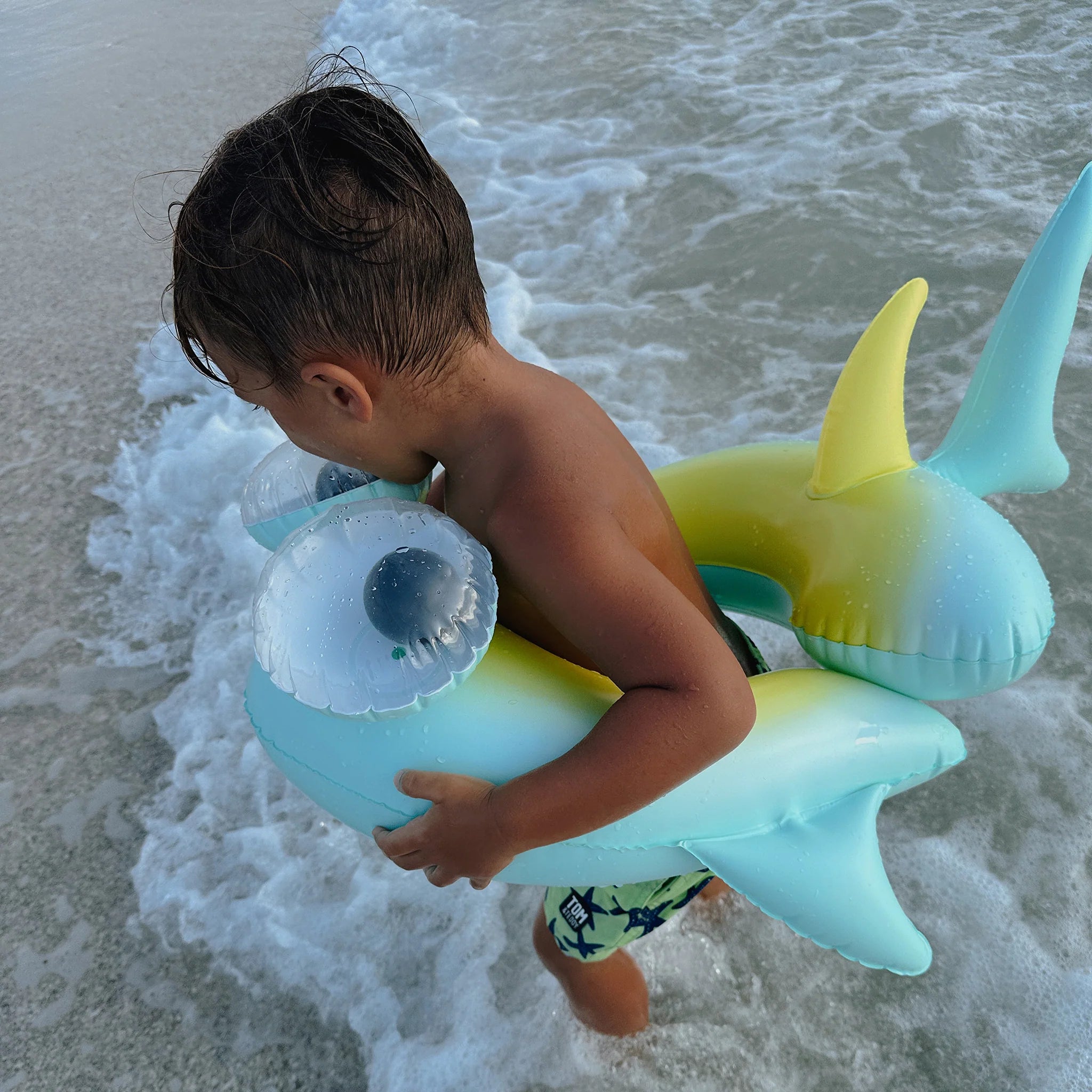 A young boy with wet hair stands at the edge of the ocean holding a SUNNYLIFE Kids Tube Pool Ring featuring Salty the Shark. He is facing away from the camera as small waves lap at his feet. Dressed in swim trunks, he appears to be playing in the water.