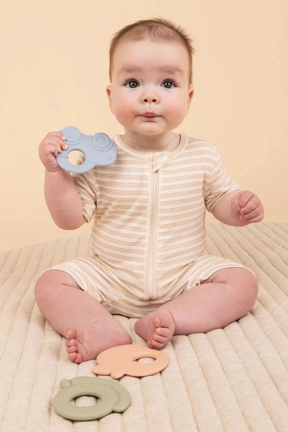 A baby in a striped onesie sits on a beige surface, holding a blue item from the KYND BABY Silicone Teething Set. Nearby, two BPA-free teething rings in pastel green and peach lie in front. The background maintains a warm, neutral tone.
