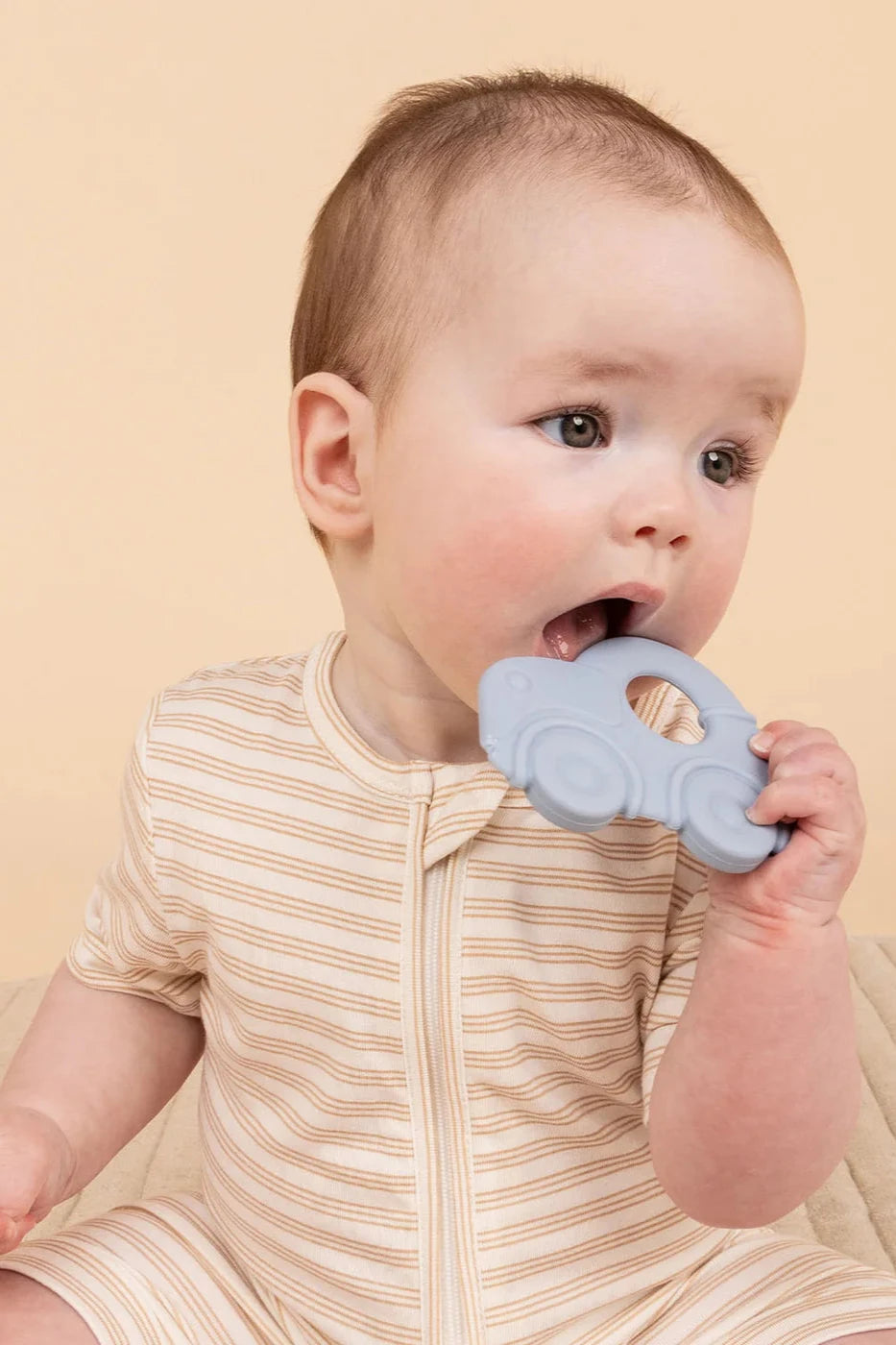 A baby in a striped onesie gently bites the blue food-grade silicone toy car from KYND BABY's Silicone Teething Set, set against a warm beige backdrop, ideal for safe exploration with this BPA-free item.