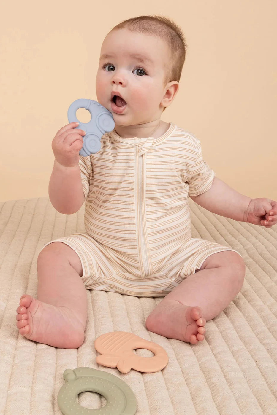 A baby in a striped onesie sits on a quilted blanket, holding the KYND BABY Silicone Teething Set to their mouth. Two other silicone teething rings from the set rest nearby on the beige background.