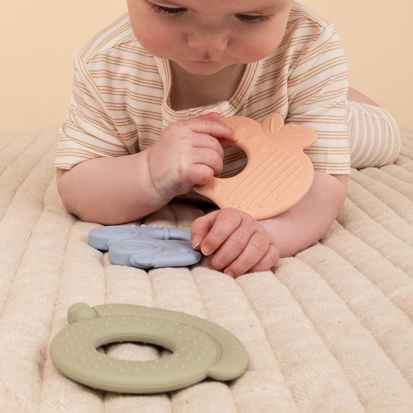 A baby in a striped onesie lies on a quilted surface, examining and holding KYND BABY's Silicone Teething Ring in BPA-Free peach. Nearby are food-grade silicone rings from the set in blue and green.