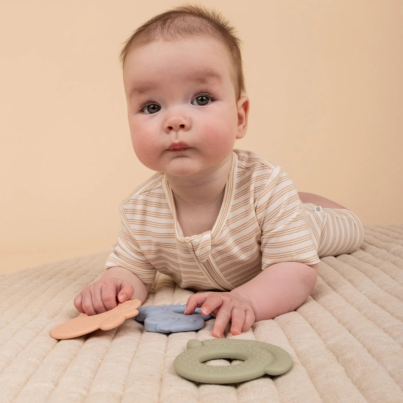 A baby in a striped onesie lies on a cushioned surface, gazing curiously forward. In front are three BPA-free teething rings from the KYND BABY Silicone Teething Set, shaped like a bear, flower, and donut.