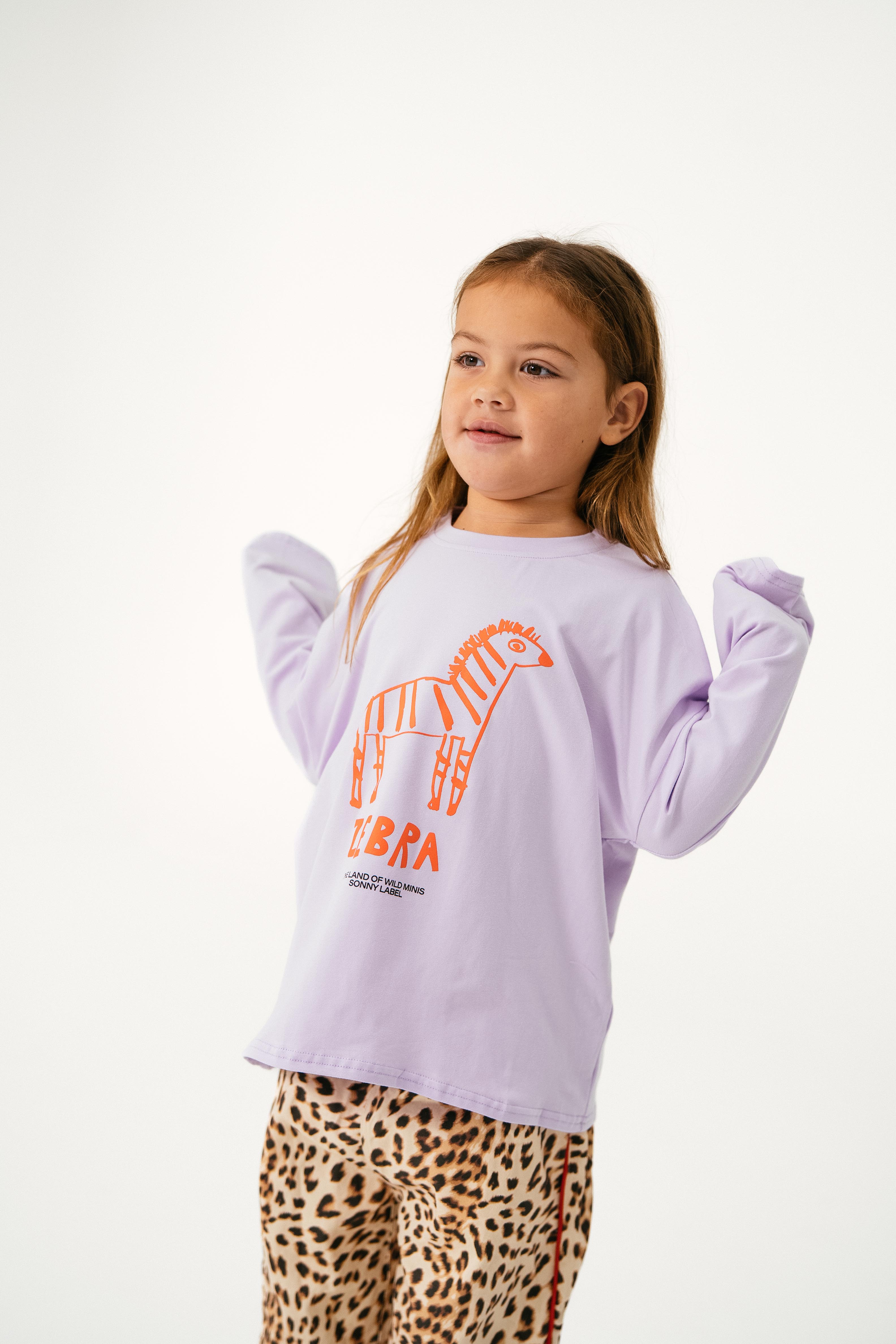A young girl with long light brown hair wears the Sonny Label - Animal Long Tee Lilac by SONNY LABEL, featuring an orange zebra graphic and "Zebra," paired with leopard print pants against a plain white background.
