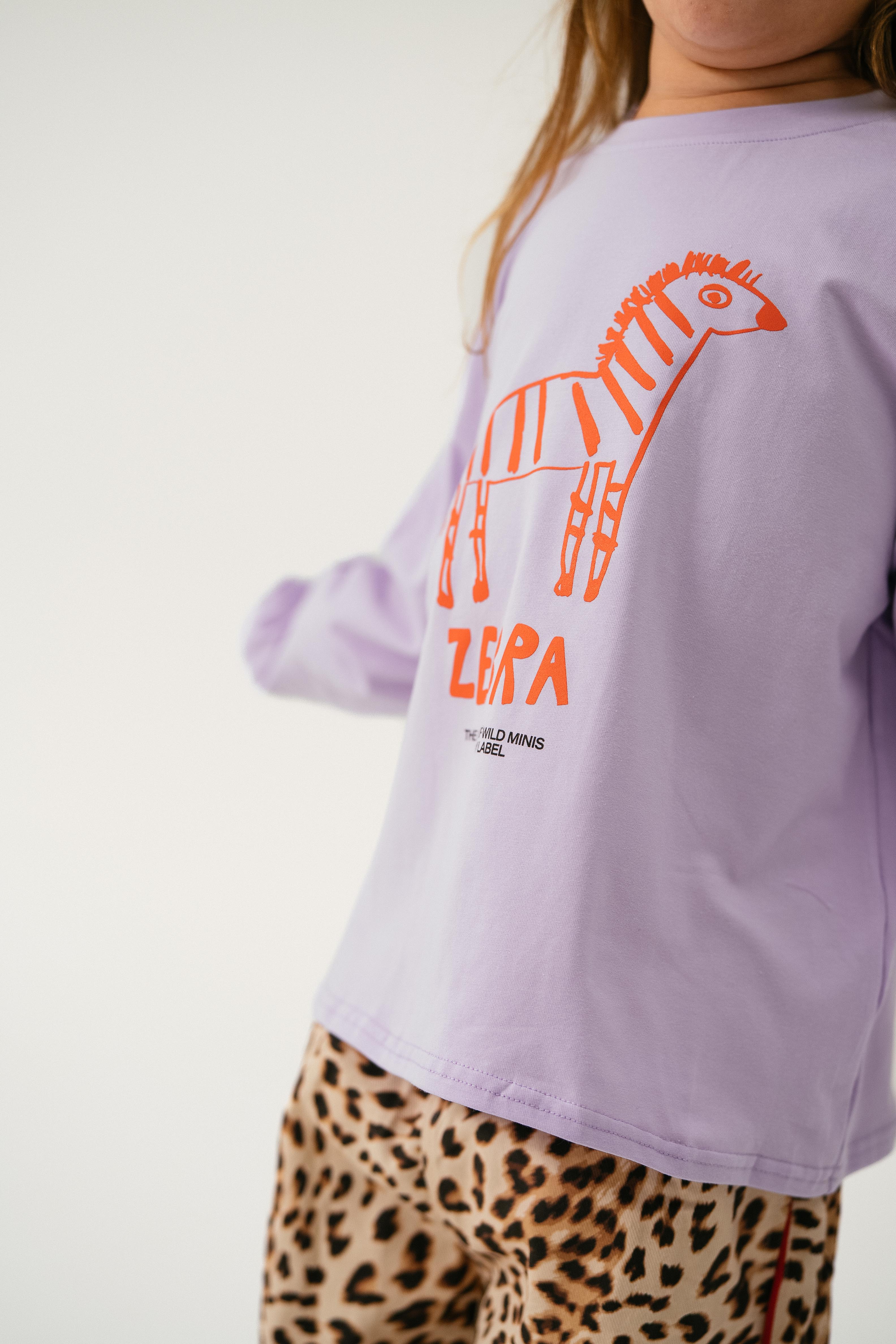 A child wears the Sonny Label - Animal Long Tee Lilac by SONNY LABEL, featuring an orange zebra graphic and “ZEBRA,” paired with leopard-print pants, standing against a plain light background.