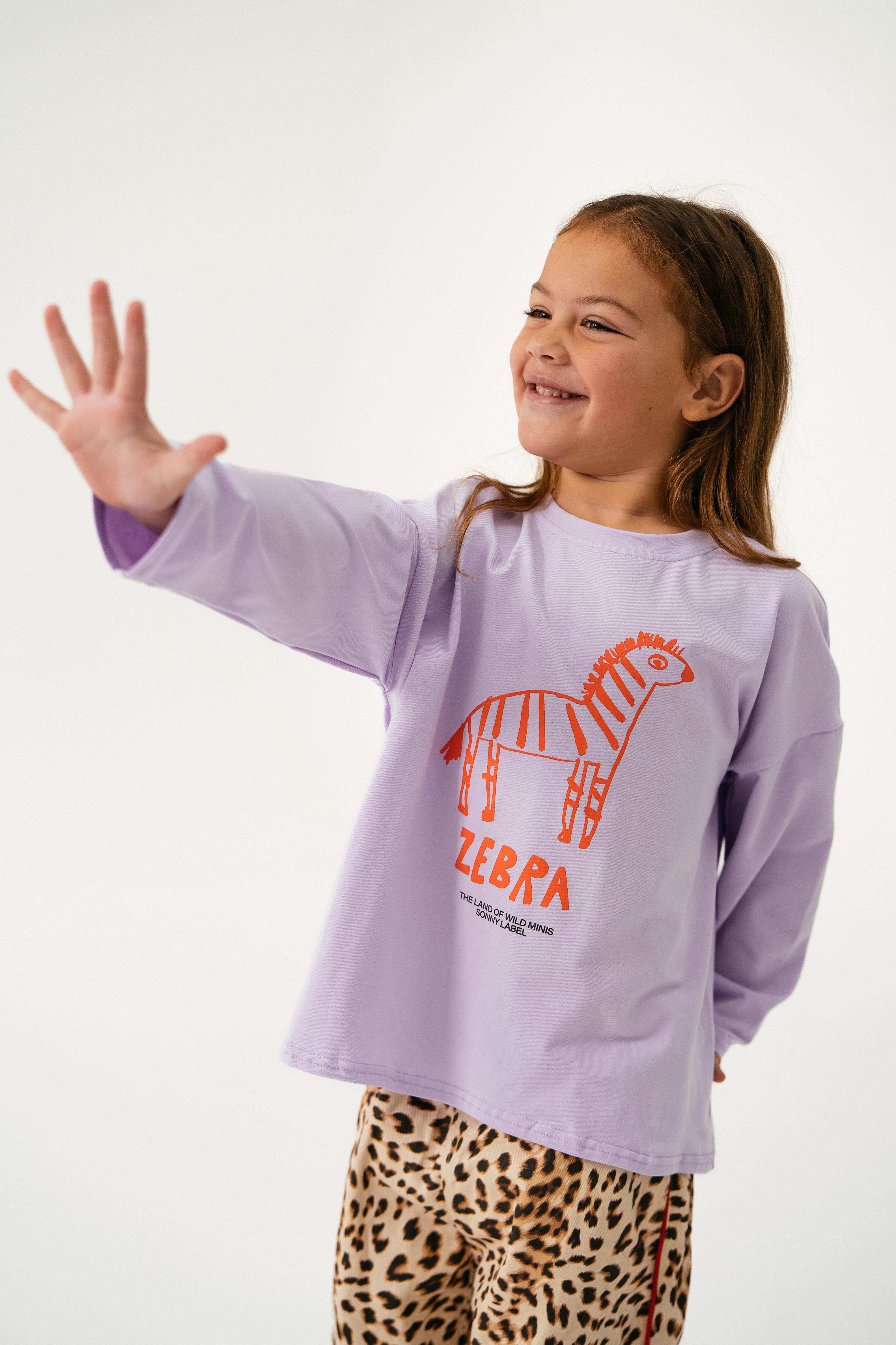 A young girl smiles and raises her hand against a white background, wearing the SONNY LABEL Sonny Label - Animal Long Tee Lilac featuring an orange zebra graphic and leopard print pants.