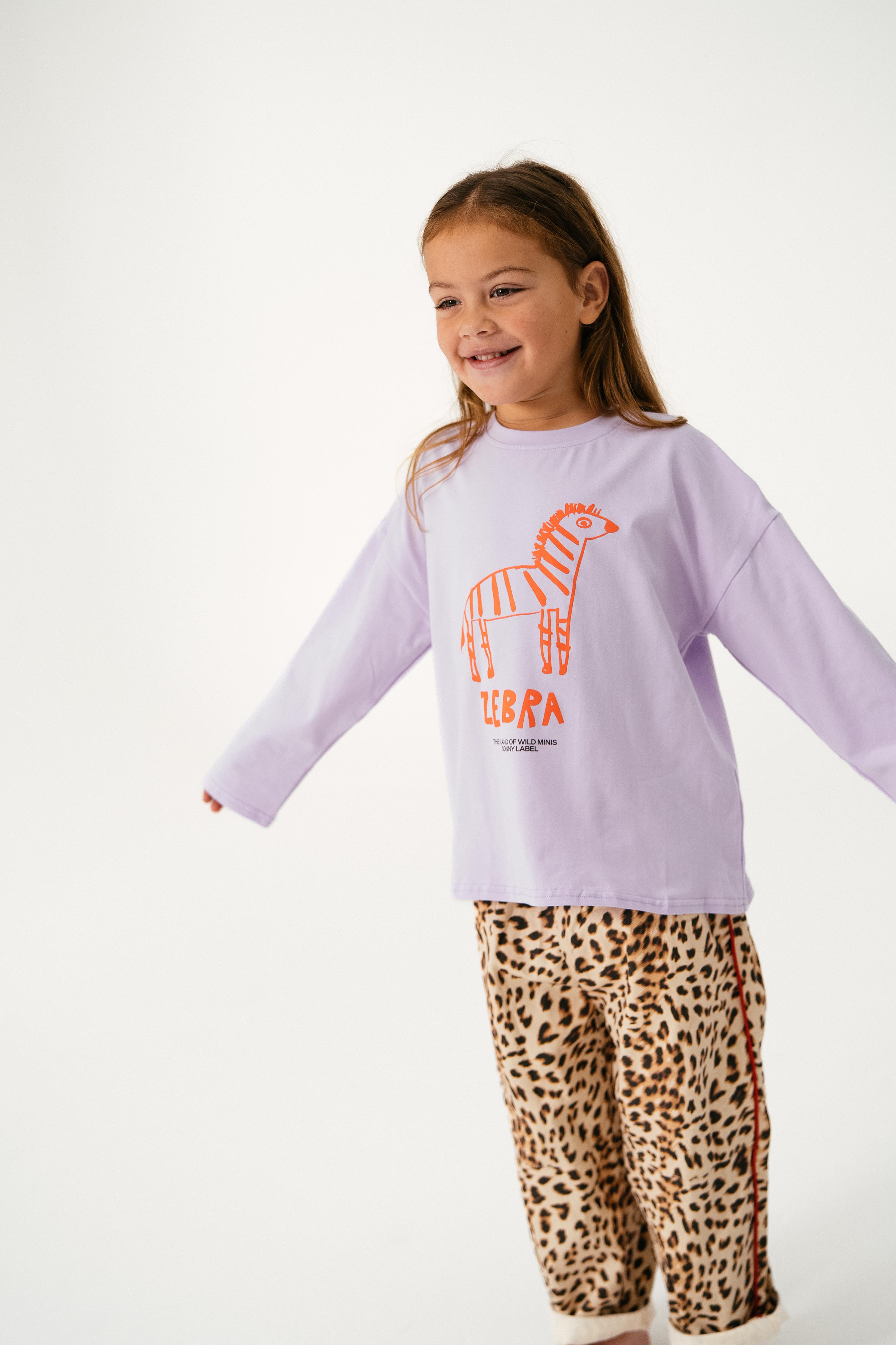 A young girl smiles with outstretched arms, wearing the SONNY LABEL Sonny Label - Animal Long Tee Lilac featuring an orange zebra graphic and text, paired with leopard print pants, set against a plain white background.