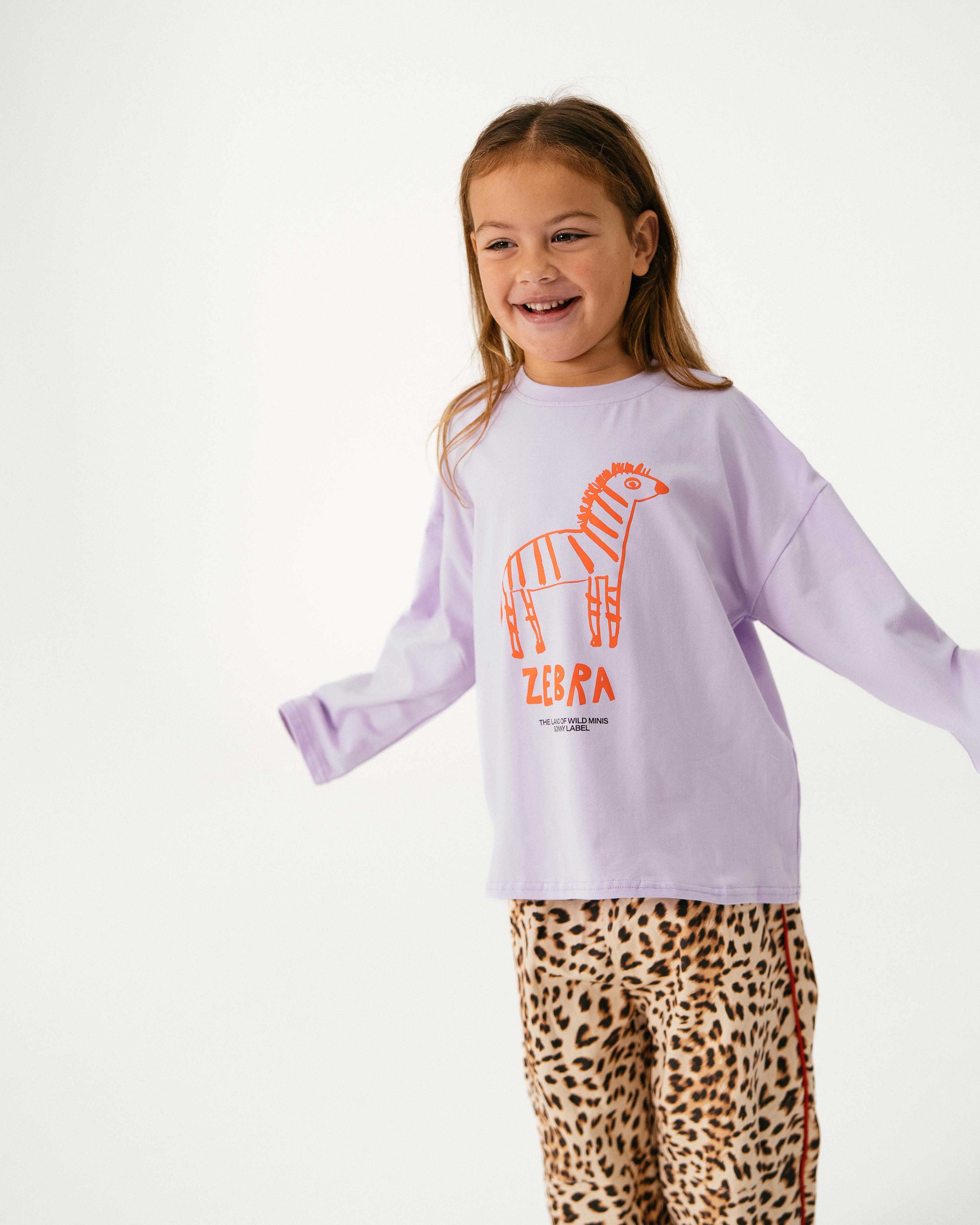 A young girl with outstretched arms smiles in the SONNY LABEL Sonny Label - Animal Long Tee Lilac, featuring an orange zebra graphic and "ZEBRA" text, paired with leopard print pants against a plain white background.