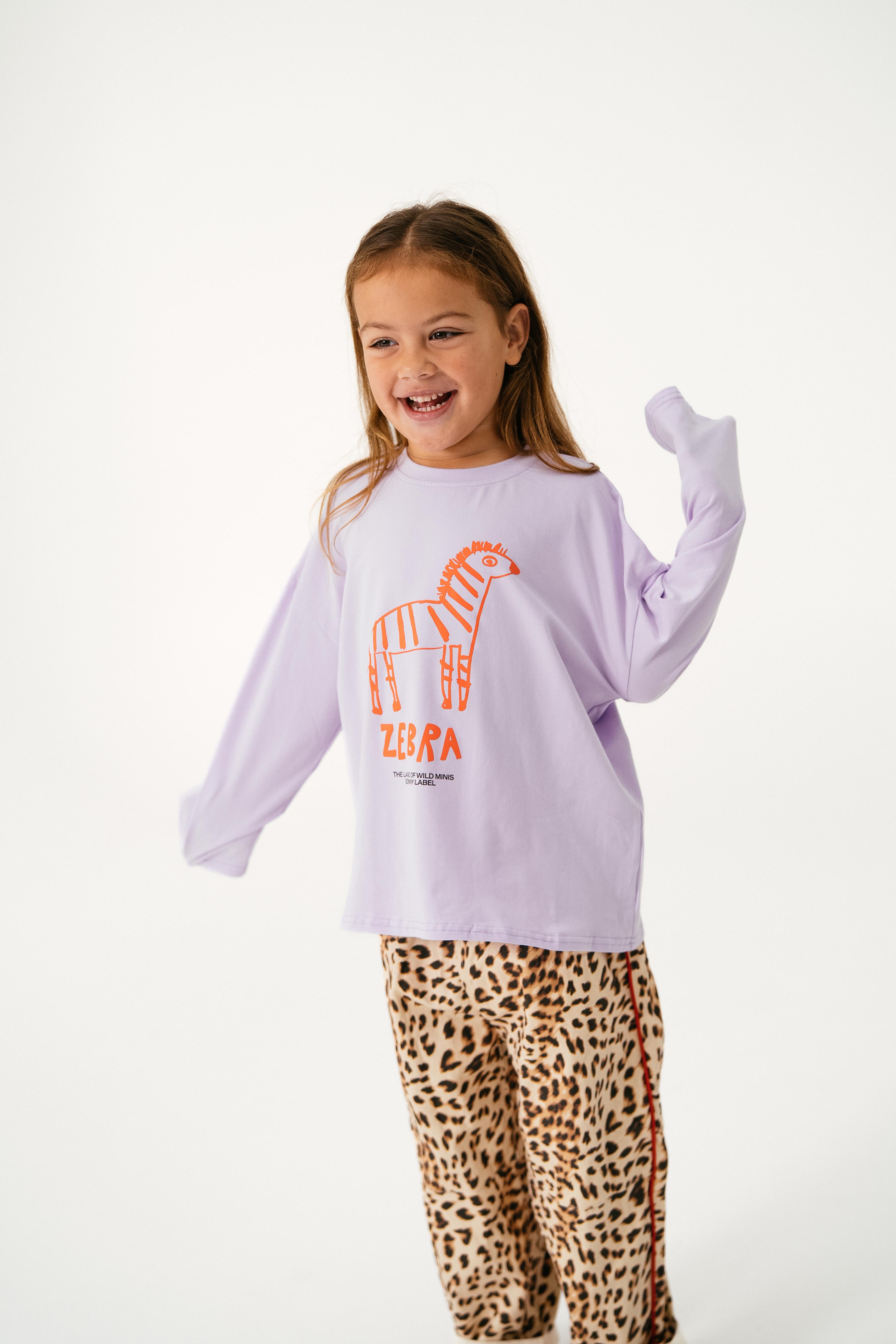 A young girl beams in the SONNY LABEL Sonny Label - Animal Long Tee Lilac, a comfy cotton-spandex top with an orange zebra graphic and "Zebra" text, paired with leopard-print pants, set against a plain white background.