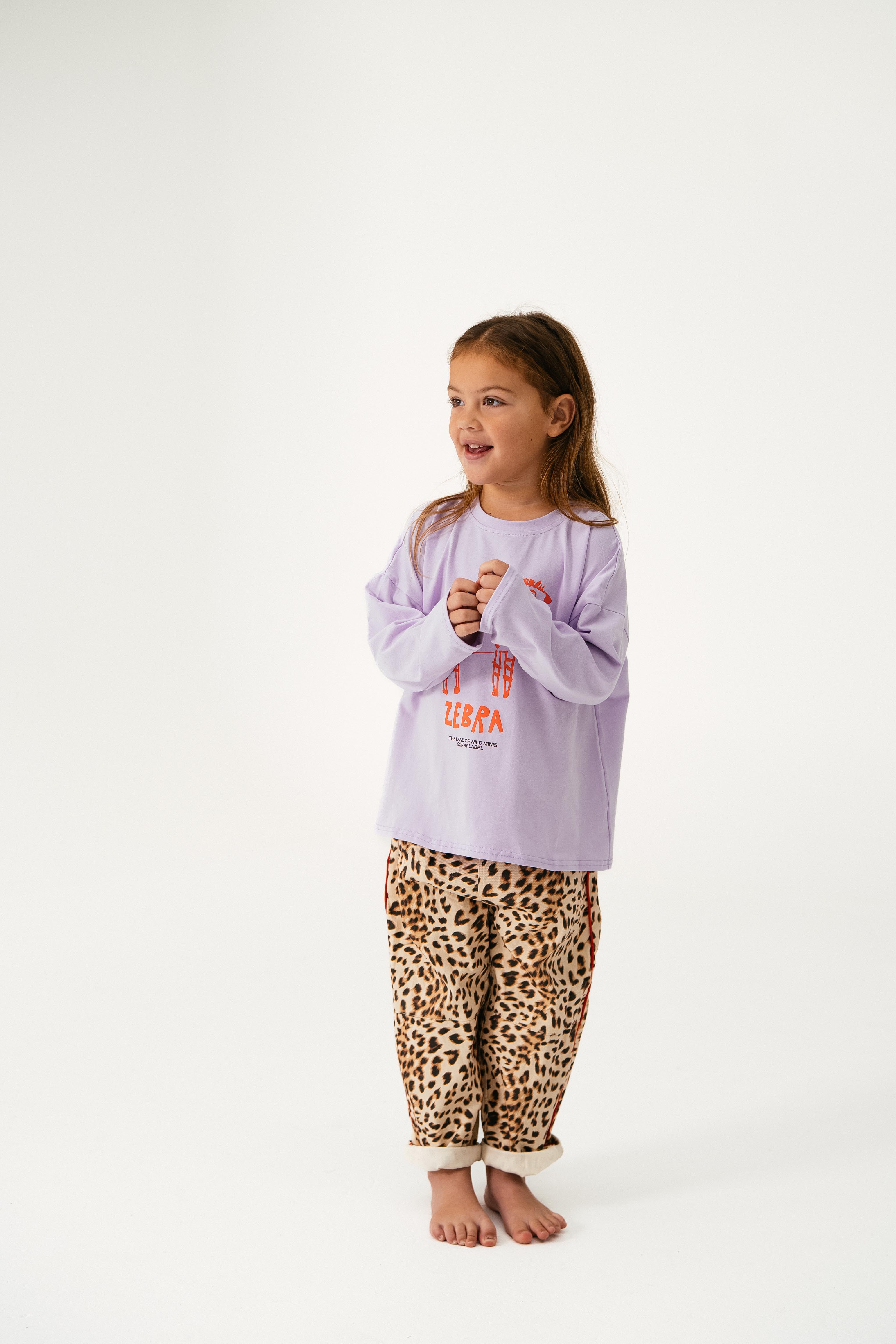A young girl stands barefoot, smiling and clasping her hands. She wears the SONNY LABEL “Animal Long Tee Lilac” with red designs and beige leopard print pants, set against a white background.