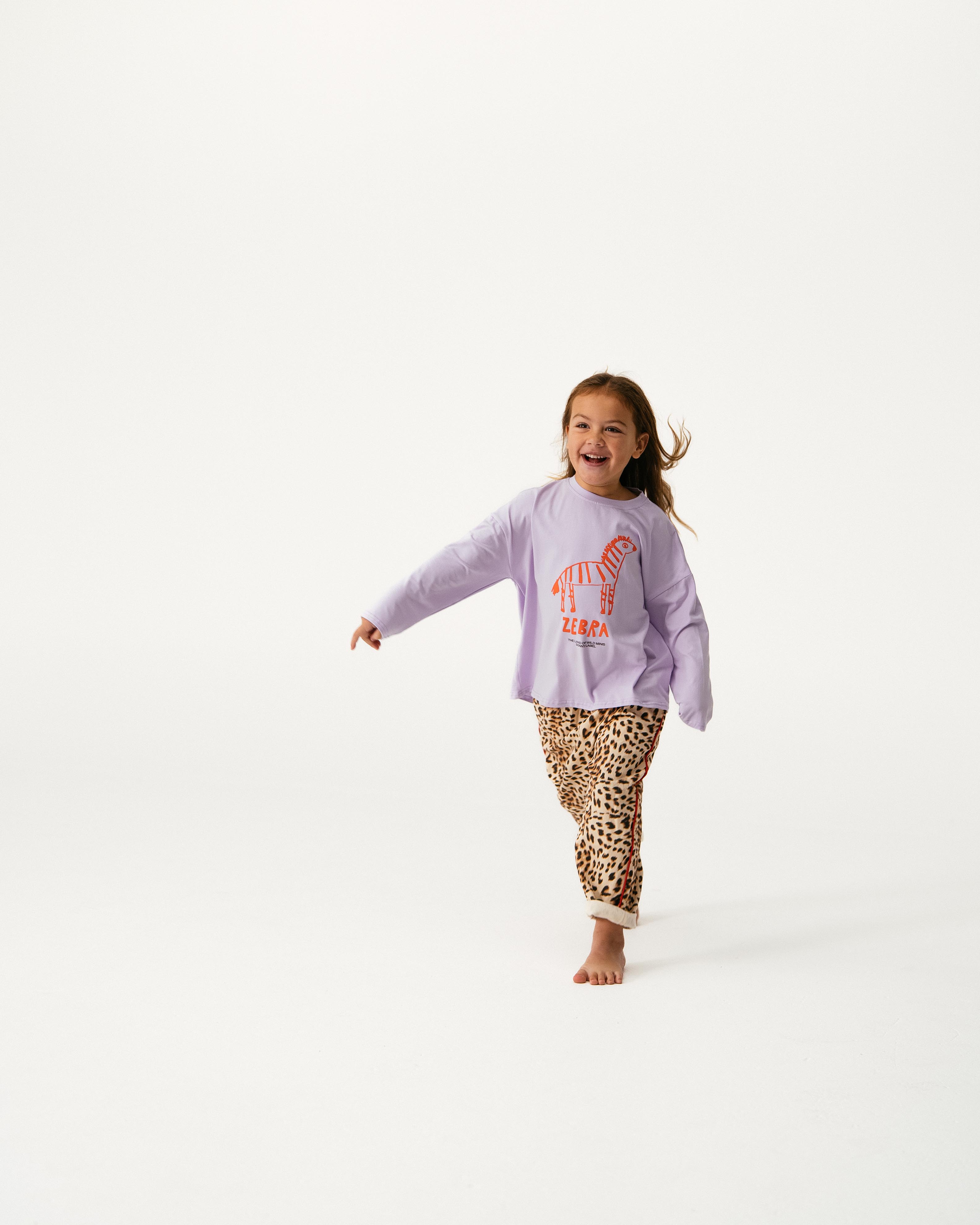 A young girl smiles as she runs barefoot on a white background, wearing the SONNY LABEL Sonny Label - Animal Long Tee Lilac with an orange zebra graphic and "ZEBRA" text, paired with leopard print pants.