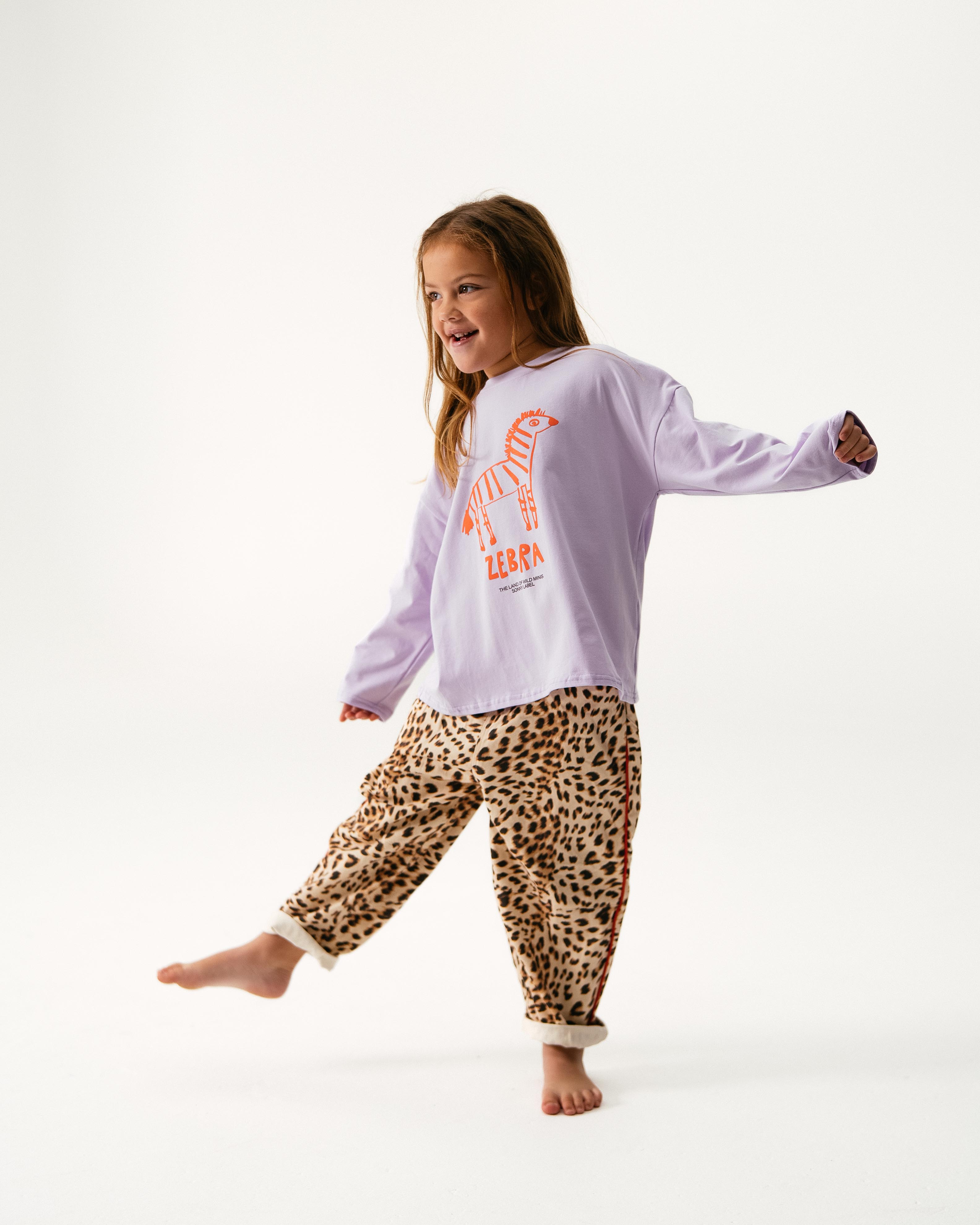 A young child stands on one foot, smiling to the side, wearing the SONNY LABEL "Animal Long Tee Lilac" with a red zebra graphic and beige leopard print pants. The background is plain white.
