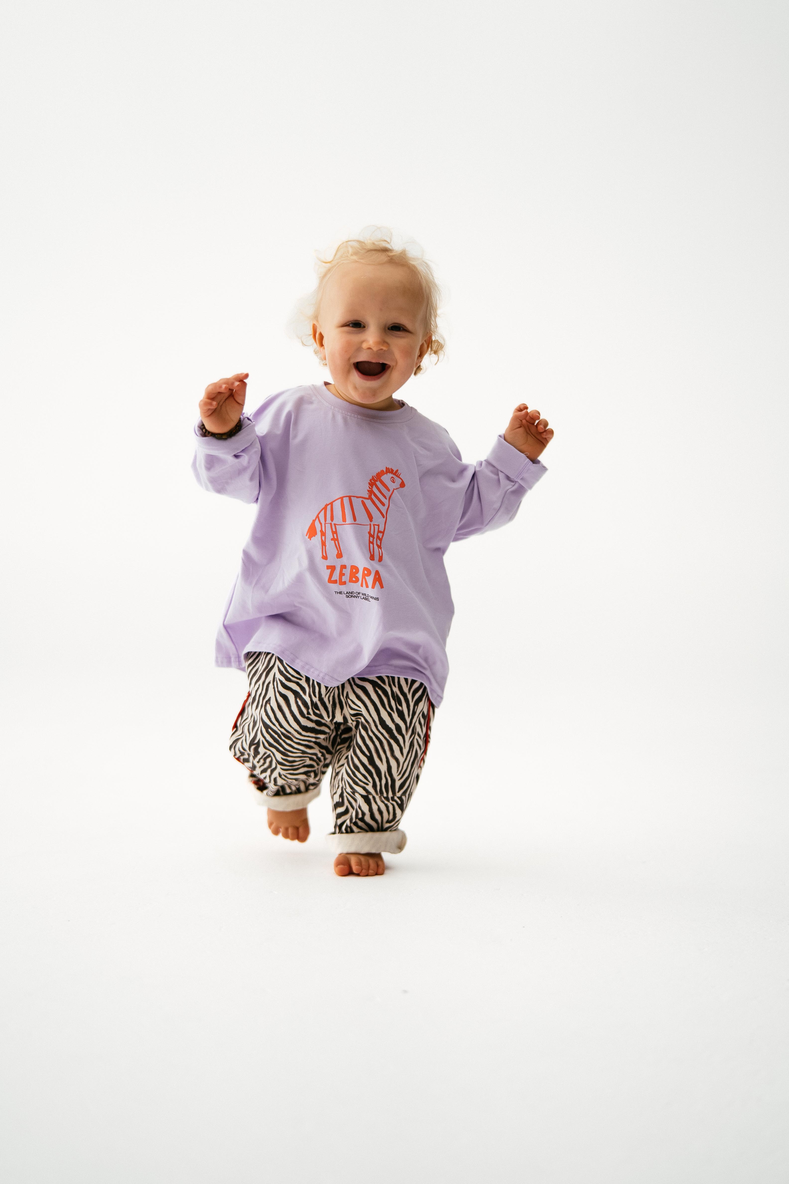 A smiling blonde toddler walks toward the camera in a SONNY LABEL Animal Long Tee Lilac featuring an orange zebra graphic and "ZEBRA," paired with black-and-white zebra print pants. The background is plain white.