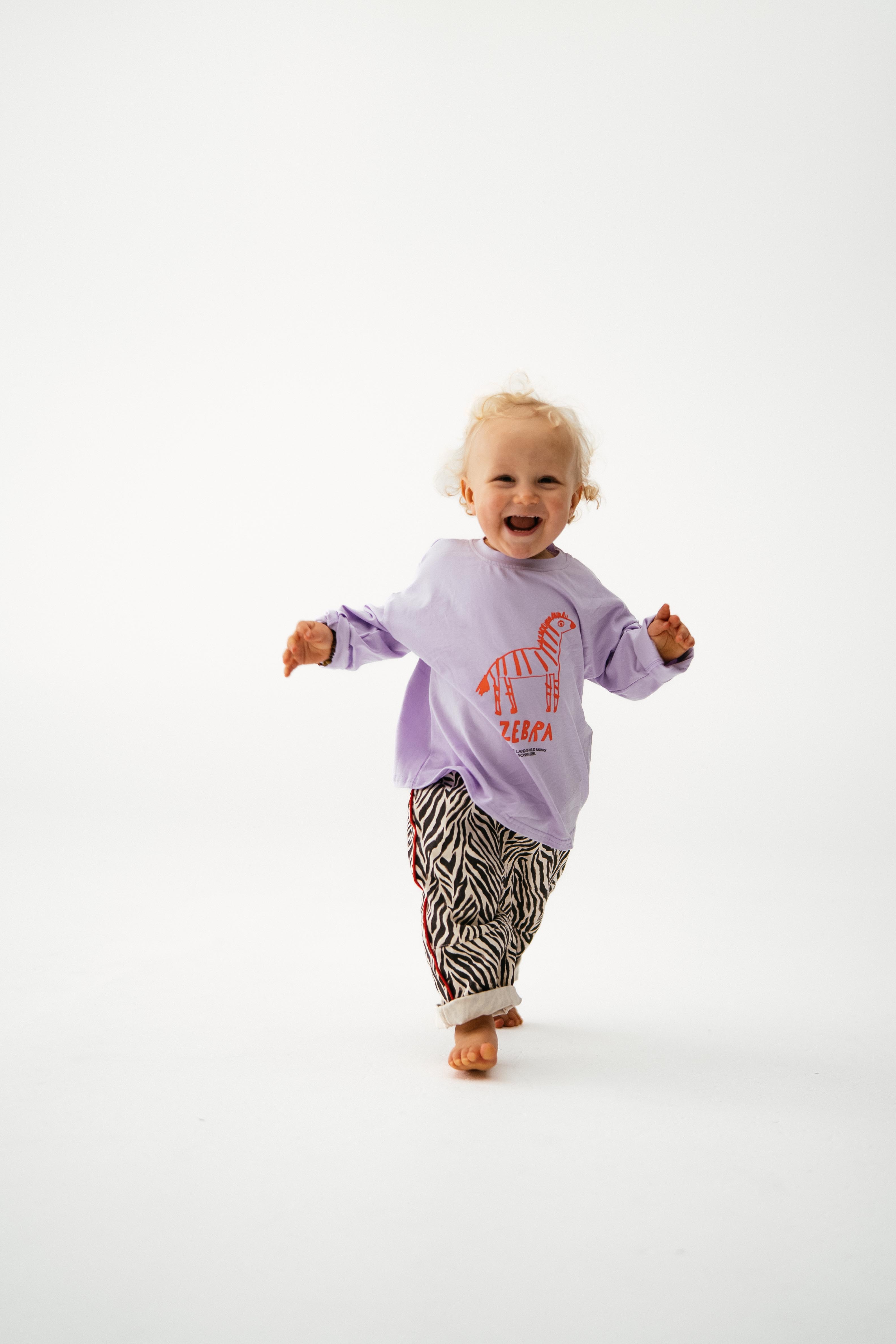 A smiling toddler with curly blonde hair runs barefoot toward the camera in the SONNY LABEL Sonny Label - Animal Long Tee Lilac, featuring an orange zebra graphic, paired with zebra-striped pants on a white background.