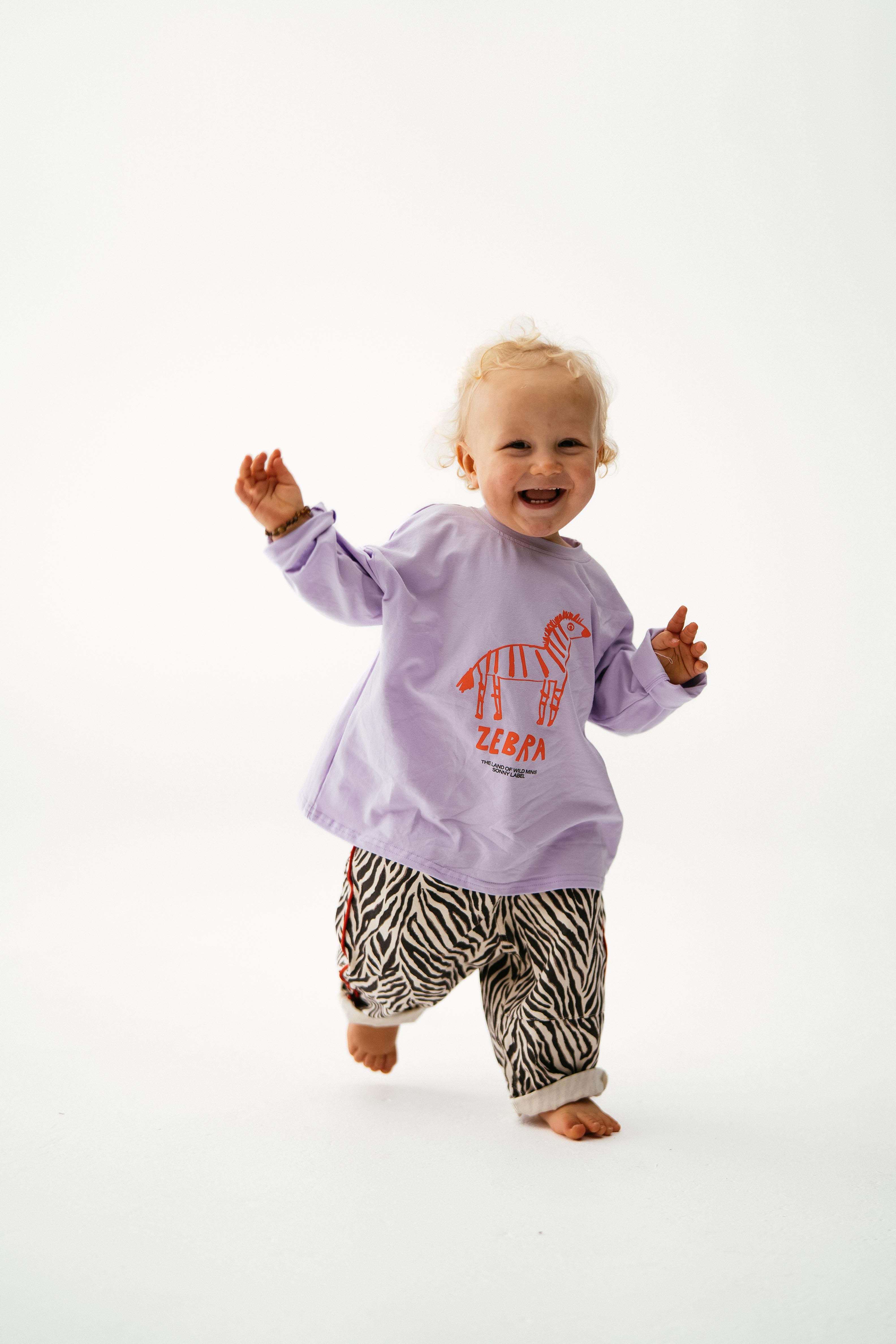 A smiling toddler with blonde hair walks barefoot in the SONNY LABEL Sonny Label - Animal Long Tee Lilac, featuring a zebra graphic and "ZEBRA" text, paired with black-and-white zebra print pants against a plain white background.