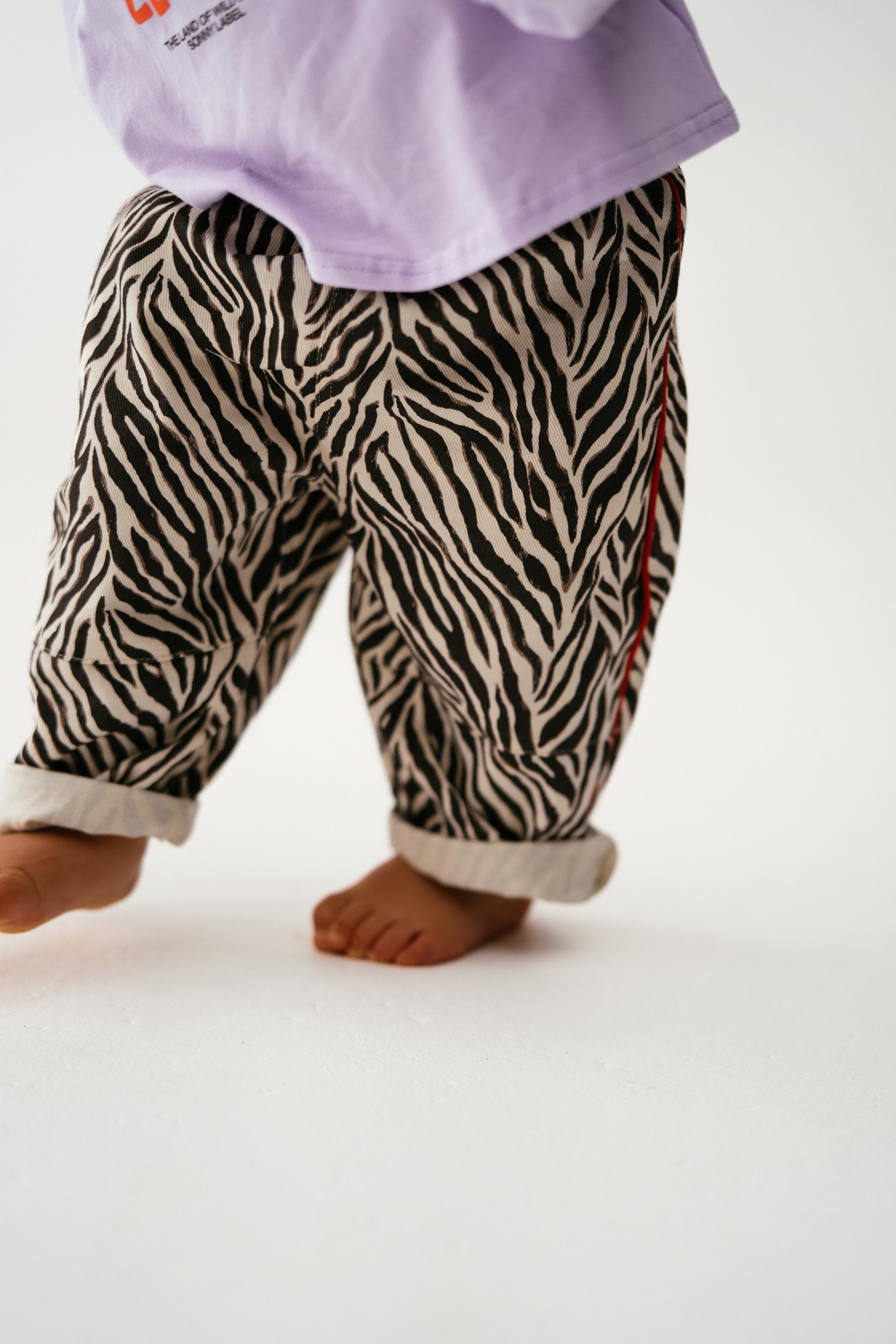 A small child stands barefoot on a white surface, wearing SONNY LABEL's "Sonny Label - Animal Twill Pants Zebra." Their upper body and face are not visible.