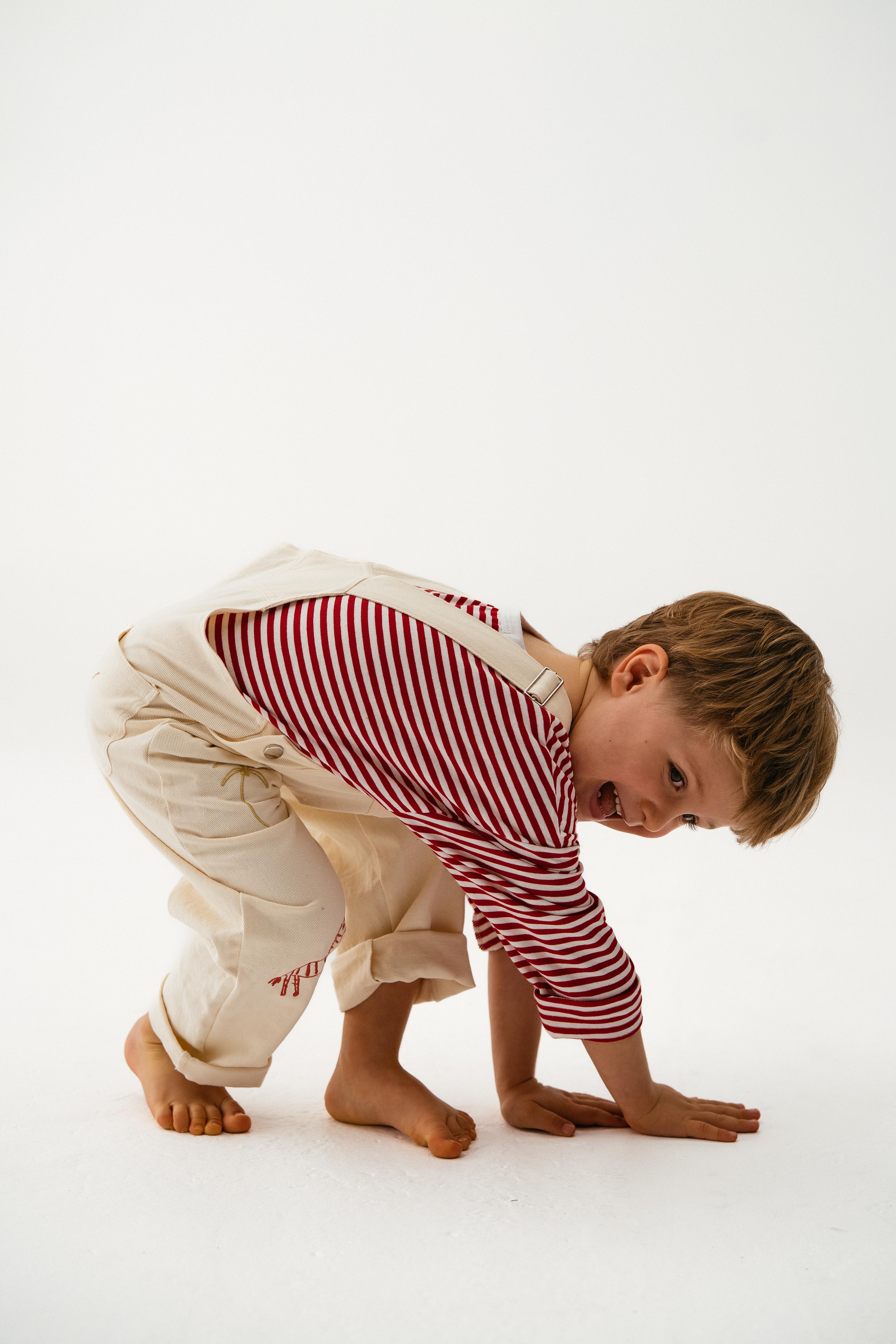 A young child in cream overalls and the SONNY LABEL Sonny Label - Striped Long Tee crouches playfully on the floor, smiling with hands touching the ground against a plain white background.