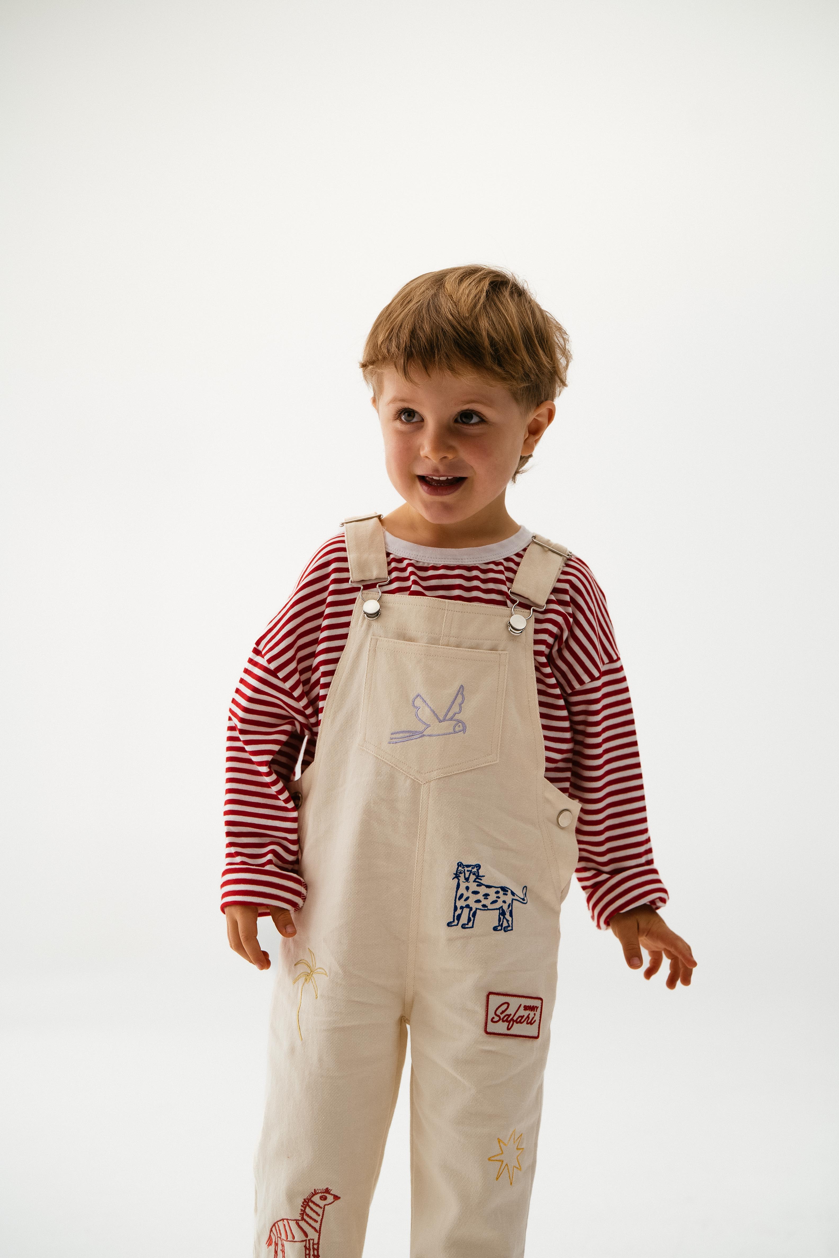 A young child with short brown hair smiles in a SONNY LABEL Sonny Label - Striped Long Tee, paired with cream overalls featuring animal patches, made from super soft cotton, standing against a plain white background.