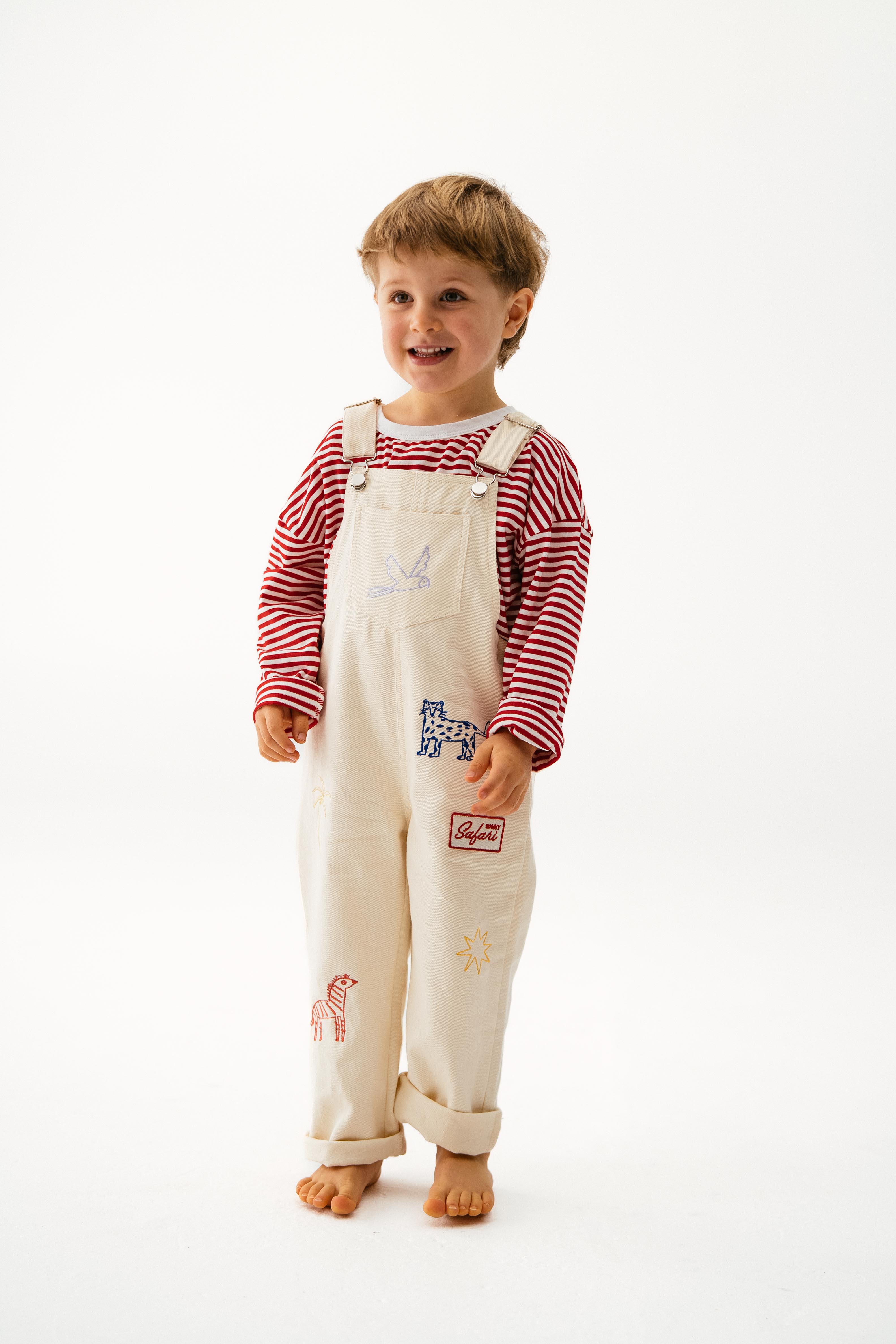 A young child smiles barefoot on a white background, wearing a SONNY LABEL Striped Long Tee in red under cream cotton overalls with fun animal and sun prints.