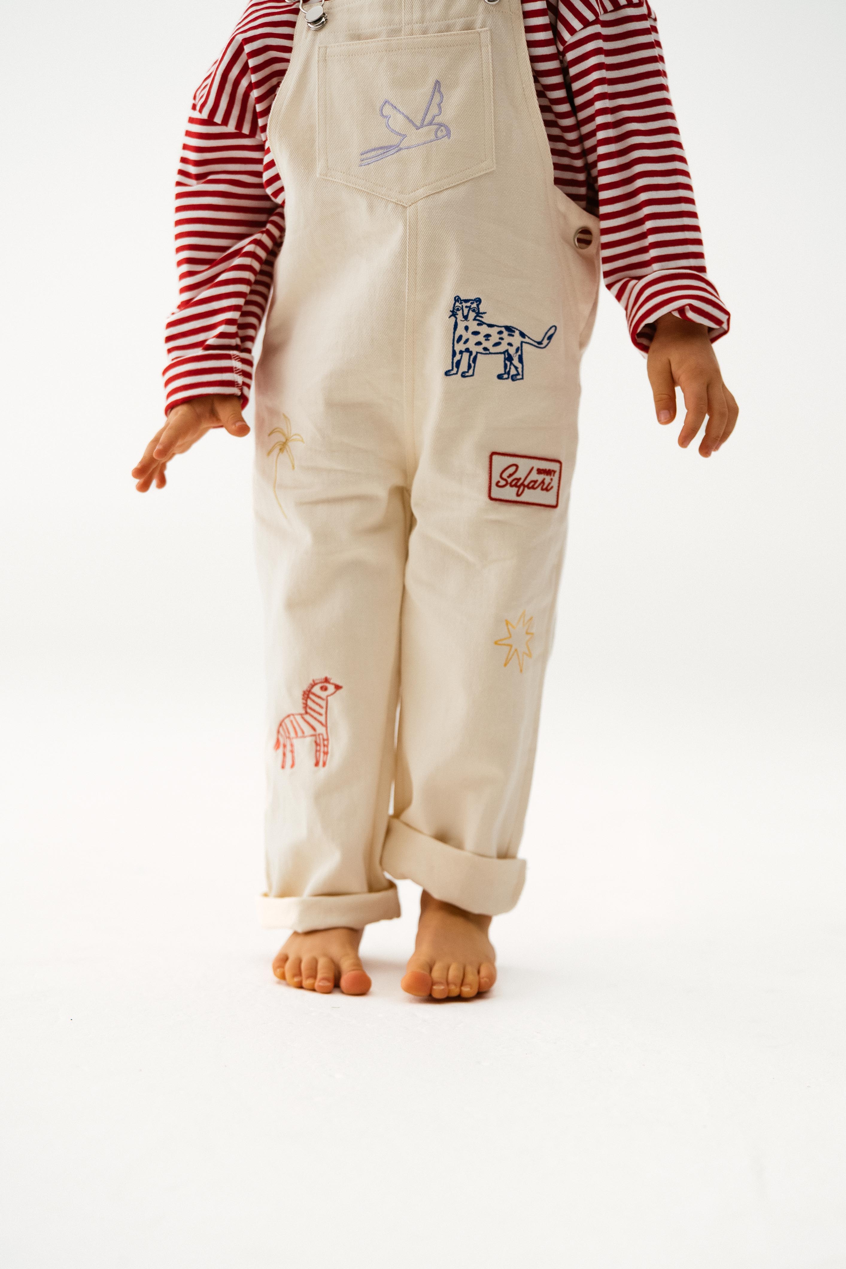 A child stands barefoot in cotton cream overalls with animal patches and "Safari" text, paired with the SONNY LABEL Sonny Label - Striped Long Tee in super soft red stripe, against a plain white background.