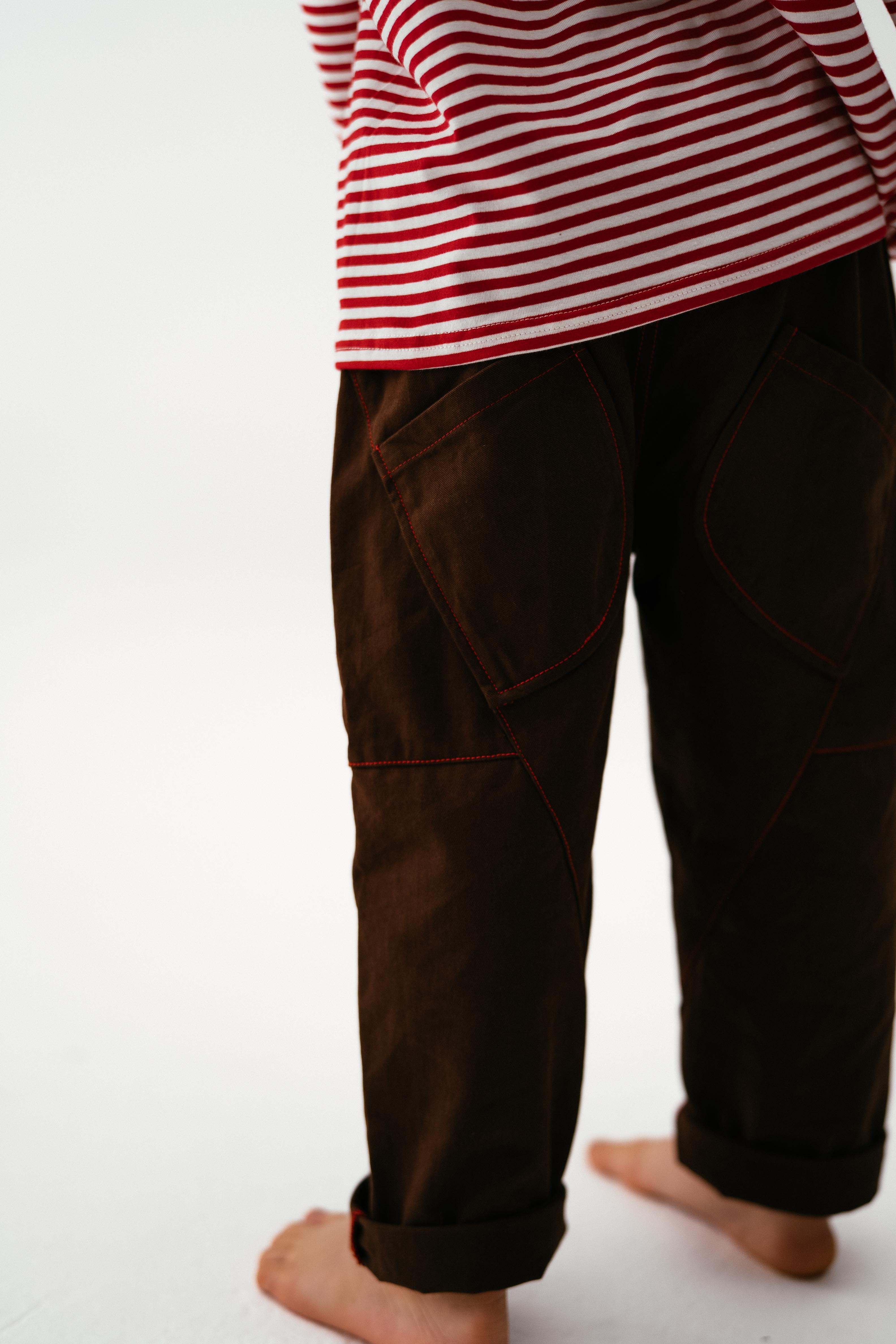 A barefoot child stands on a white surface, wearing SONNY LABEL's Sonny Label Cargo Pants in dark brown cotton twill with red stitching. The child's upper body and face are not visible.