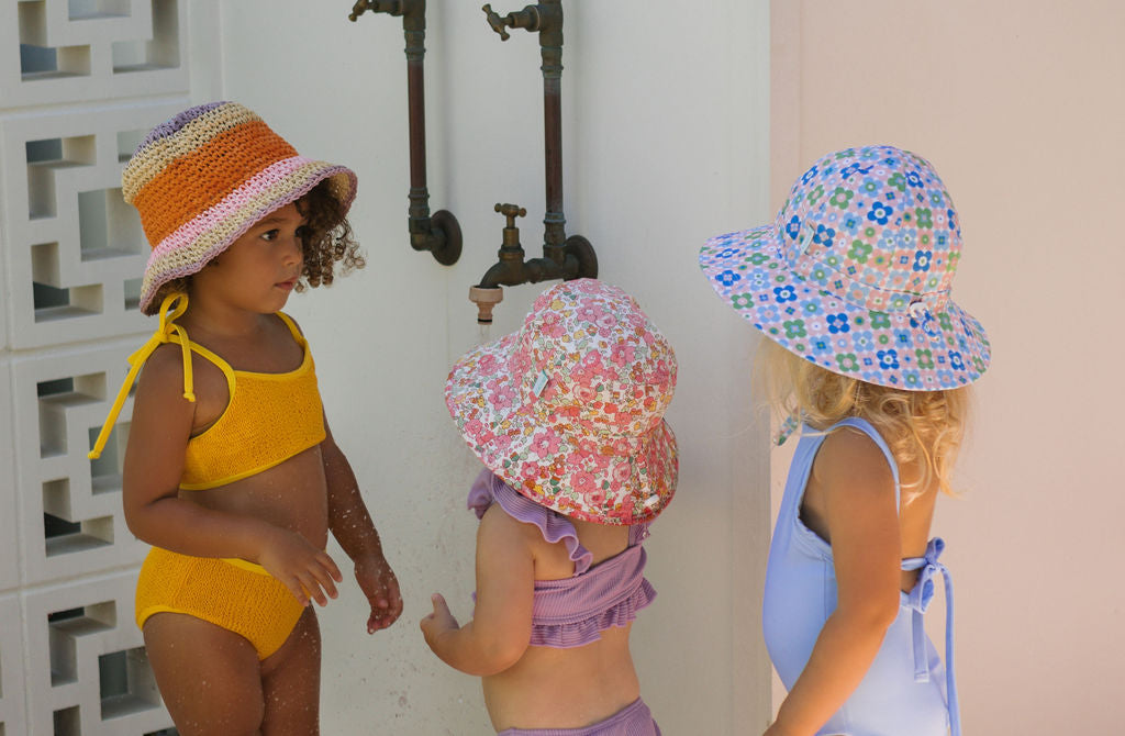 Three young children wearing ACORN KIDS Acorn Kids - Astrid Crochet Straw Hats and colorful swimsuits stand outdoors by a wall with water pipes, chatting and playing together in the sunlight.