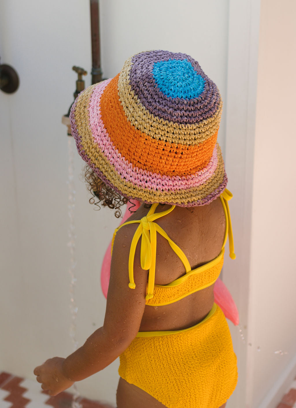A young child in a yellow swimsuit wears the Acorn Kids - Astrid Crochet Straw Hat by ACORN KIDS, standing under an outdoor shower with water running, facing away from the camera.
