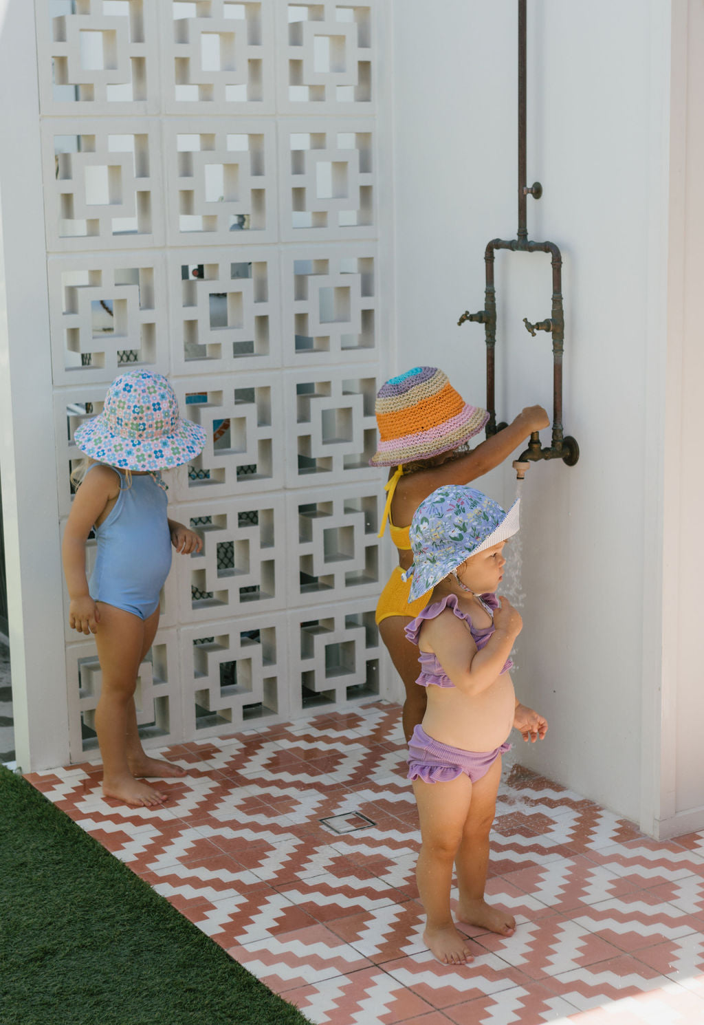 Three young children in colorful swimsuits stand on patterned tiles near a white wall; one turns an outdoor shower handle while the others watch. One child wears the ACORN KIDS Astrid Crochet Straw Hat.