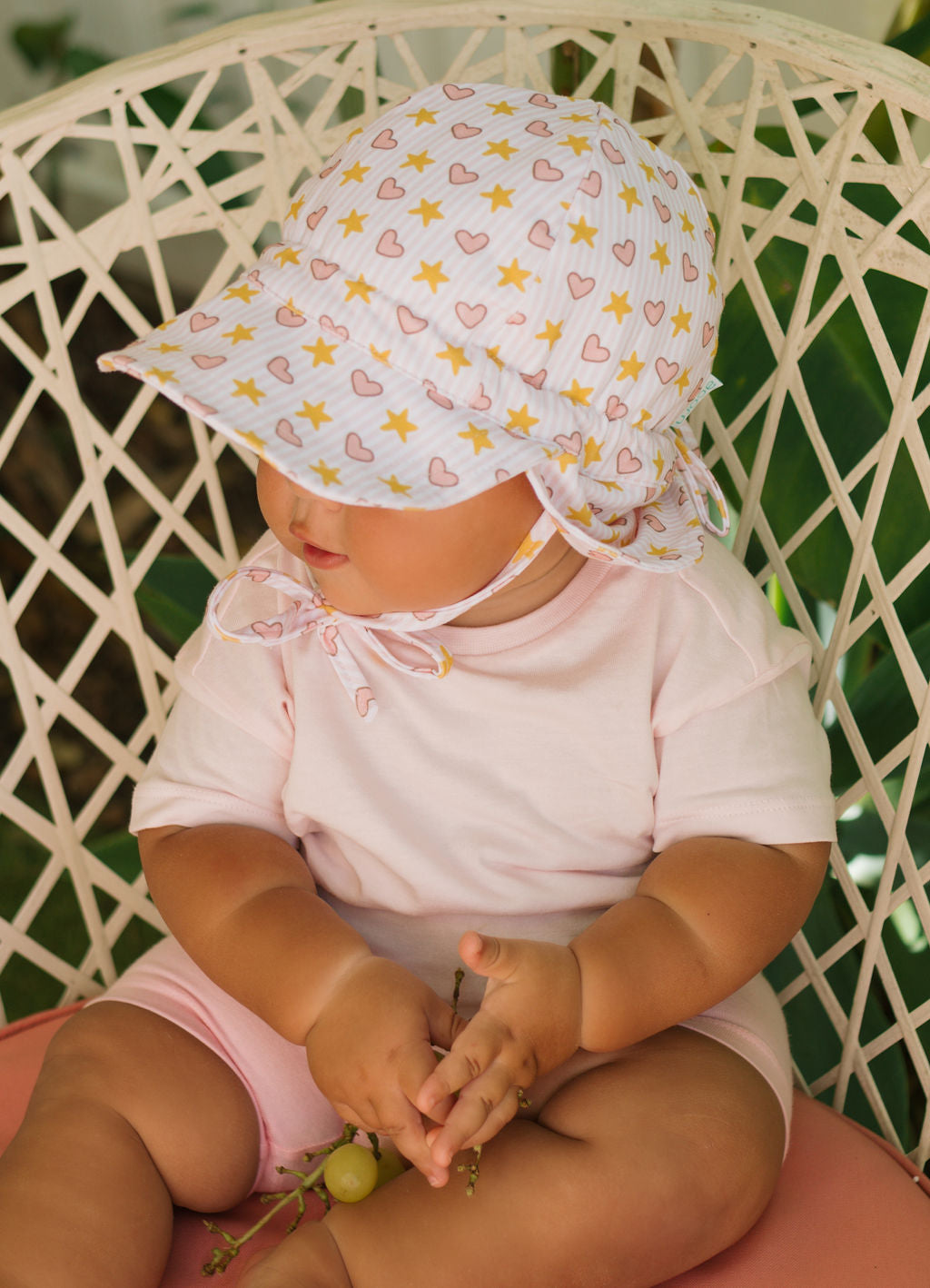 A baby in a pink outfit and an ACORN KIDS Acorn Kids - Love and Stars Flap Hat, offering UPF50+ sun protection, sits on a chair holding green grapes under lush green leaves. The cotton jersey hat features yellow stars and pink hearts.