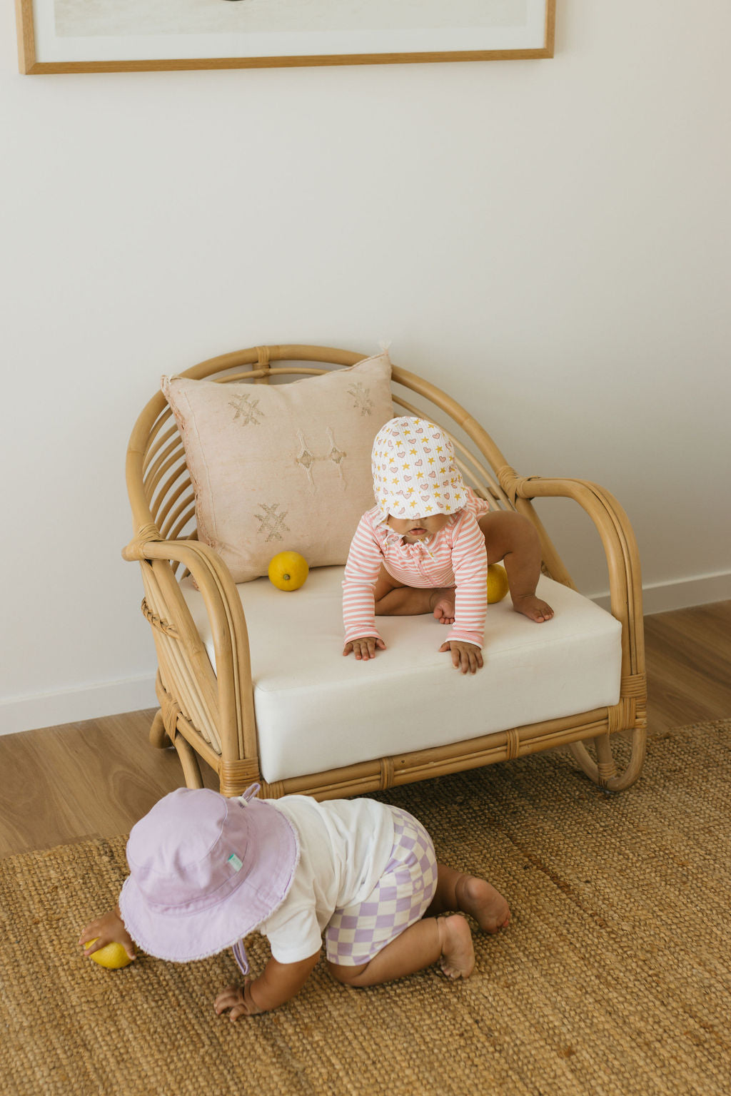 Two babies wear ACORN KIDS - Love and Stars Flap Hats as they play indoors; one sits on a rattan chair holding a lemon, while the other crawls on a woven rug, surrounded by light walls and wooden floors.