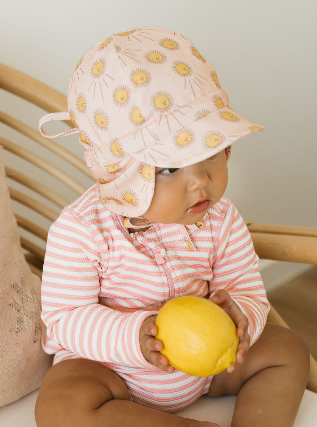A baby in a soft pink and white cotton jersey outfit and an ACORN KIDS Acorn Kids - Sunshine Flap Hat with yellow sun patterns sits on a wooden chair, holding a bright yellow lemon.