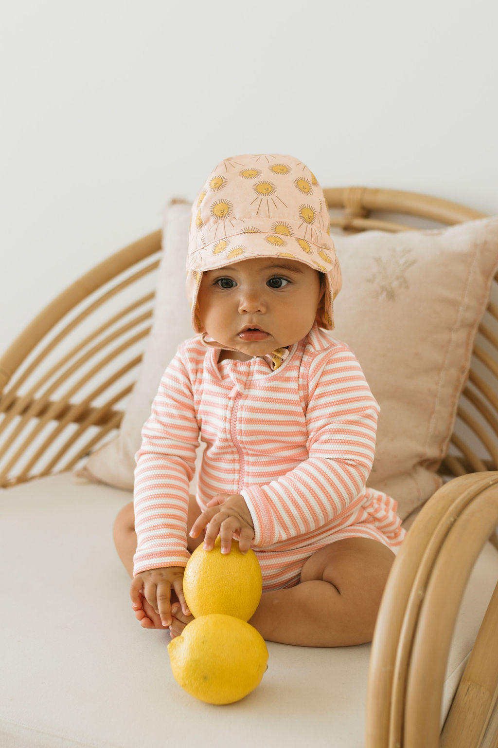 A baby wearing a pink and white striped cotton outfit and the ACORN KIDS Acorn Kids - Sunshine Flap Hat with UPF50+ sun protection sits on a rattan chair, holding two yellow lemons in their lap.