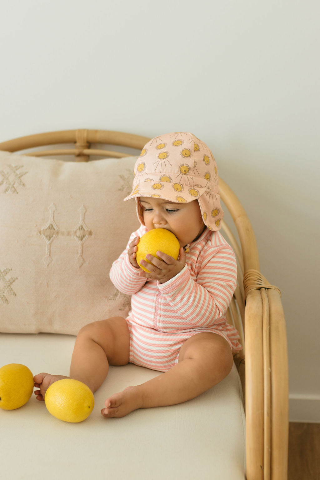 A baby in an ACORN KIDS Sunshine Flap Hat sits on a rattan chair, holding a lemon with two more beside them. The baby wears a striped cotton onesie, with a beige embroidered cushion behind.