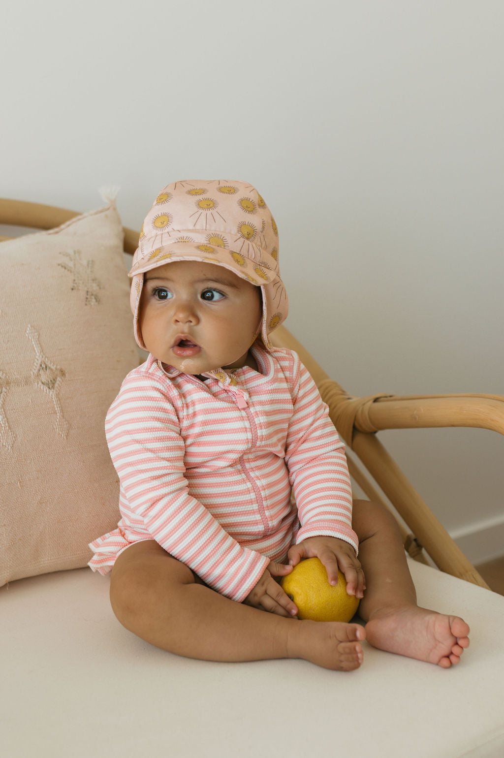A baby wears the ACORN KIDS Acorn Kids - Sunshine Flap Hat with yellow flowers and a pink-and-white striped cotton outfit, while sitting on a cushioned chair and holding a lemon, with a beige pillow behind.