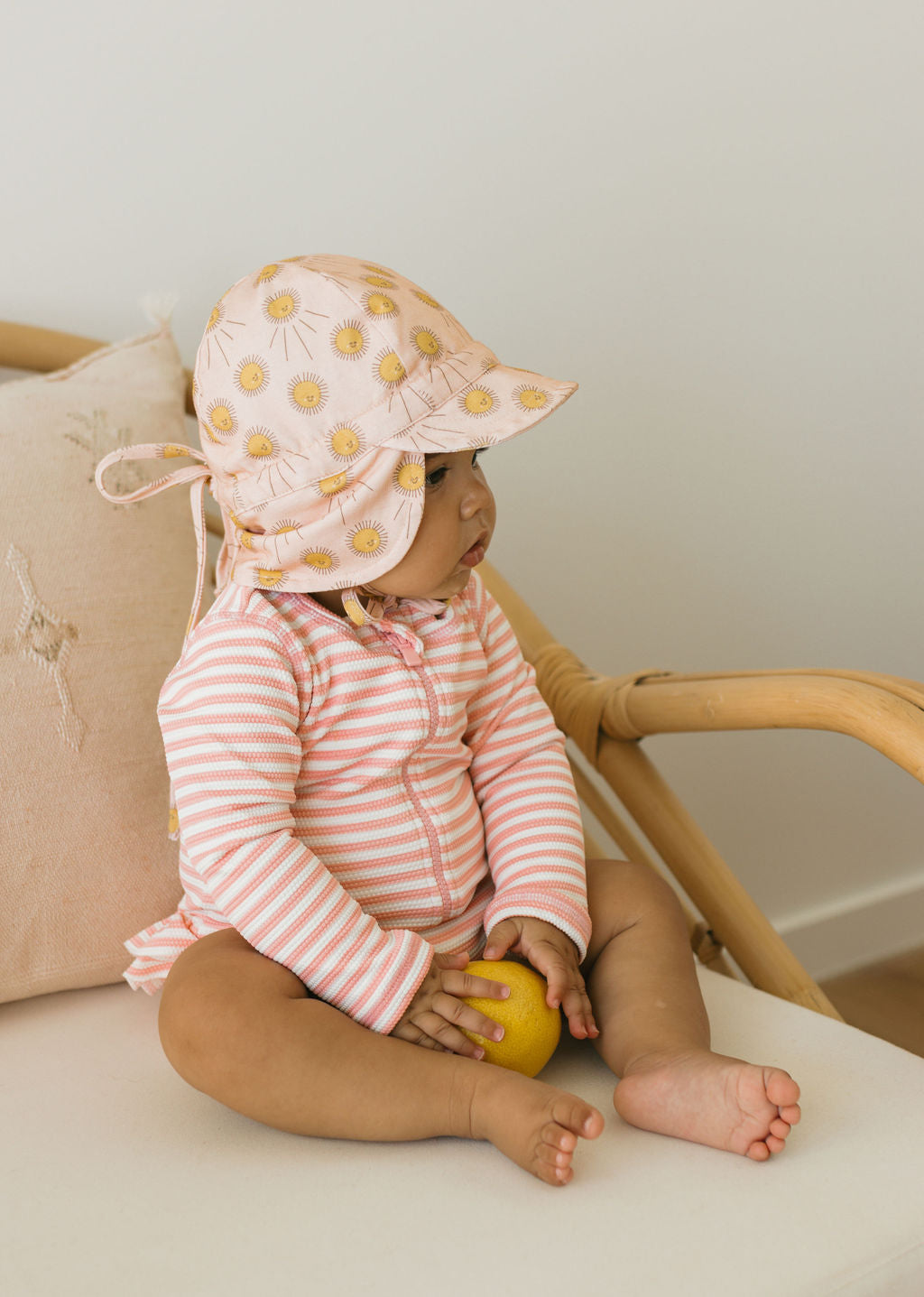 A baby in a pink and white cotton jersey outfit wears the ACORN KIDS Sunshine Flap Hat, featuring yellow flowers, while sitting on a light chair, holding a lemon and looking to the side.