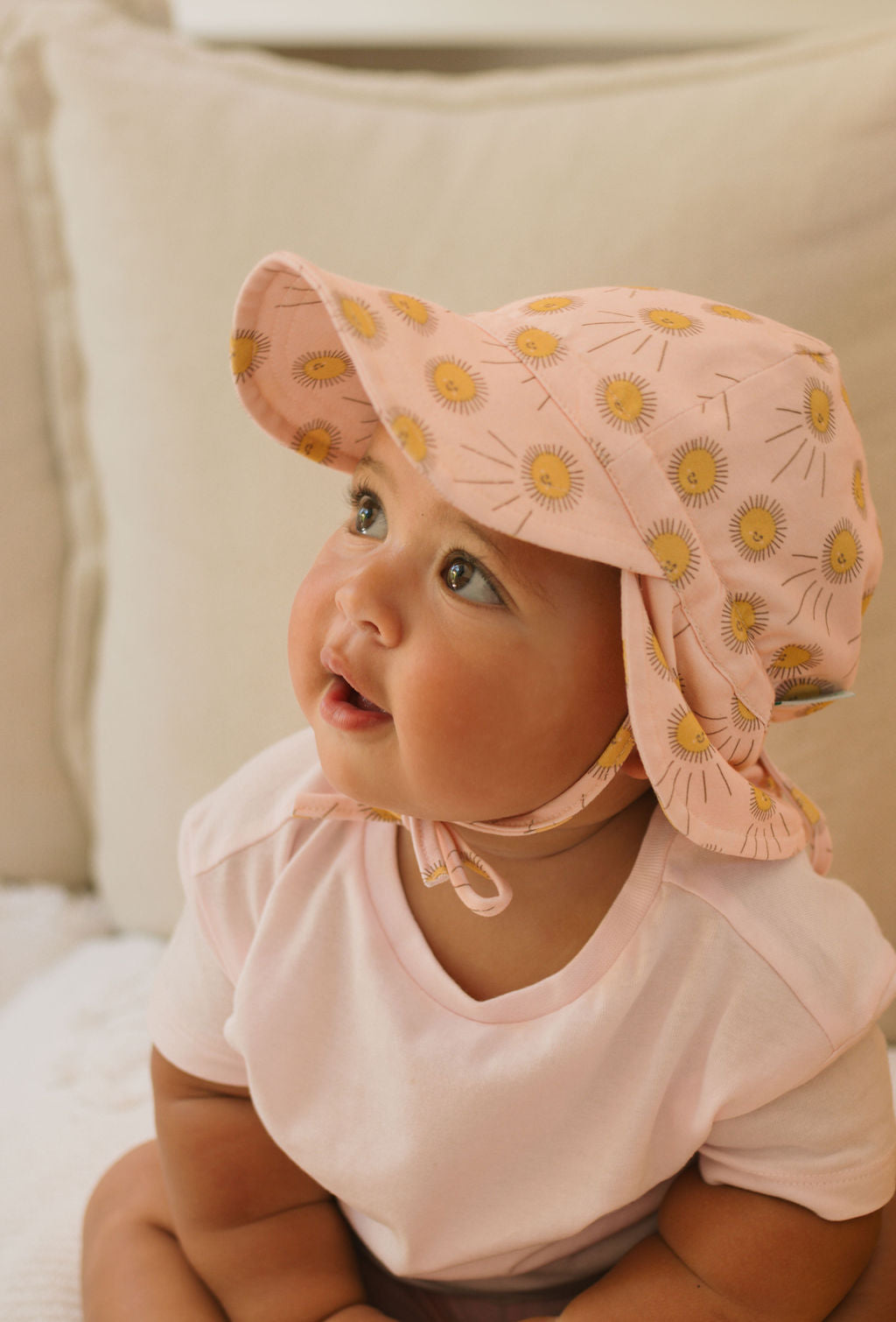 A baby wears the ACORN KIDS Acorn Kids - Sunshine Flap Hat, a pink cap with a yellow floral pattern and UPF50+ sun protection, paired with a soft cotton jersey shirt, and looks up indoors with curiosity.